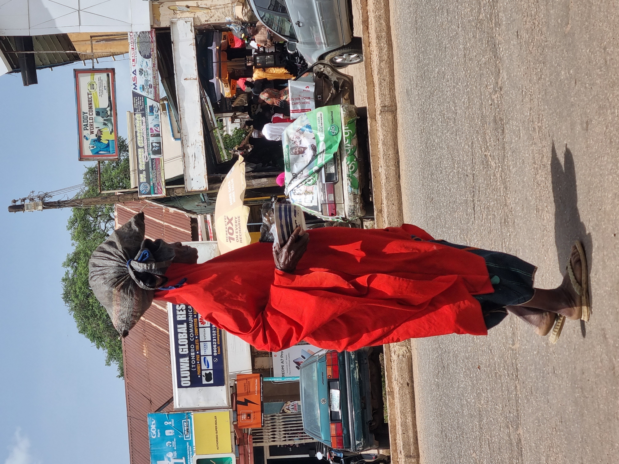 A man in a red robe and wrapped head covering walks on a paved street in Jos, Nigeria. He holds an object in his hands. Behind him, a green Volkswagen and a silver car are parked along the curb. Buildings line the street, adorned with signs advertising "Paddy World Connect," "Oluwa Global Resources," and Airtel. A sign in green and white promotes "Glo Unlimited" data plans. Other signs indicate vendors for phones, tablets, and computer accessories. The environment appears to be a bustling commercial area.