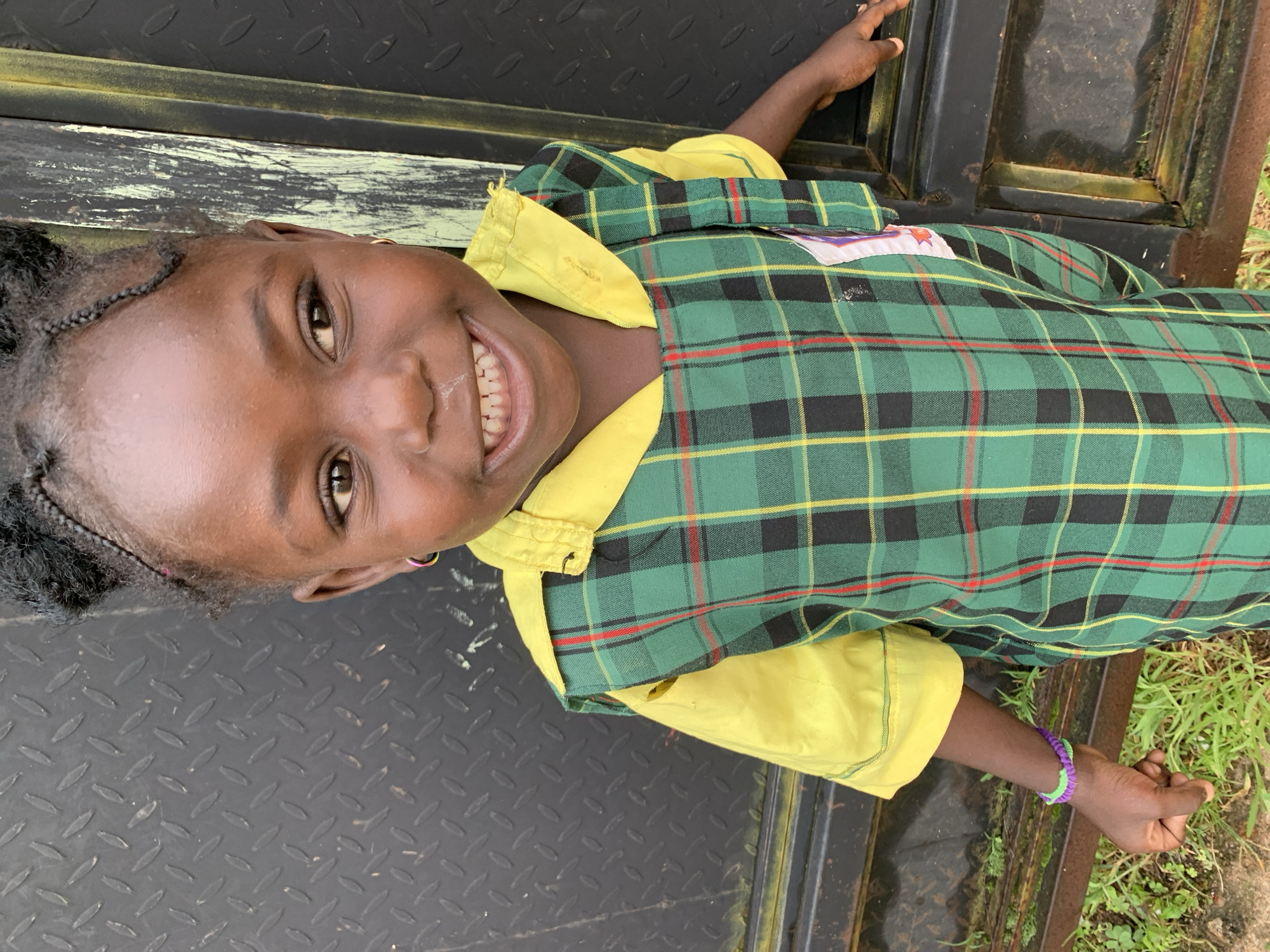 A young Black girl, appearing to be of primary school age, is captured in a close-up, eye-level outdoor portrait. She is smiling broadly, revealing her teeth, and her gaze is directed slightly off-camera to her right. Her dark hair is styled in two braided buns atop her head. She wears a school uniform consisting of a yellow collared shirt with a green, black, and red plaid jumper over it. A purple and green beaded bracelet adorns her left wrist. Her left hand is positioned in the lower left of the frame, appearing slightly clenched, while her right hand is visible in the lower right, resting on a dark, textured metal surface. The background consists of this metal surface, which appears to be a gate or door, with some green foliage visible at the bottom. The lighting suggests it is daytime. The image is likely taken in Jos, Nigeria, given the context of a school uniform.