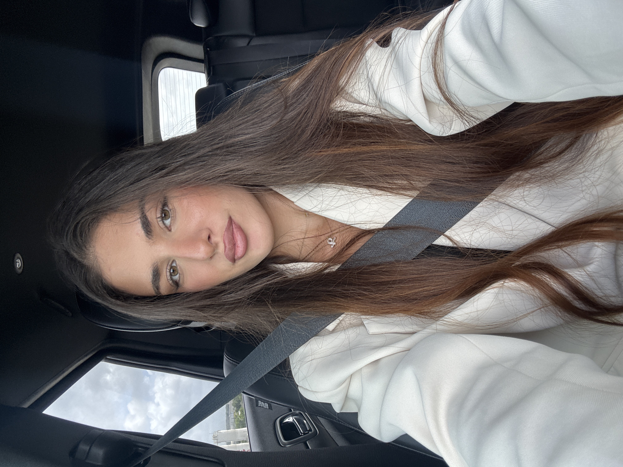 A woman is sitting in the driver's seat of a vehicle. She is wearing a white blazer and a seatbelt. The scene is in North Miami Beach, Florida, United States of America. The sky is visible through the sunroof.