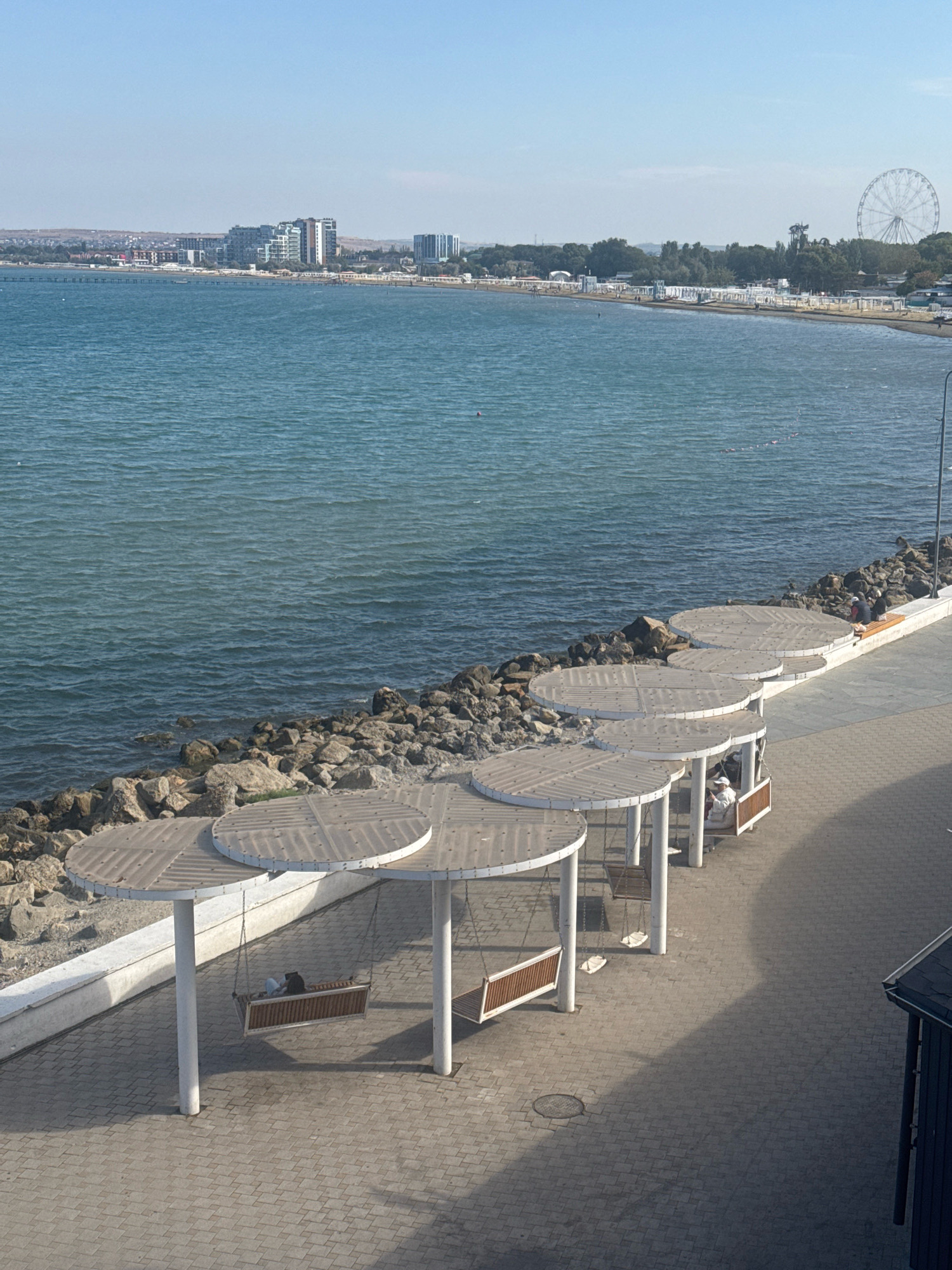 This image depicts a scenic view of the Anapa embankment in Anapa, Russia, featuring a waterfront promenade overlooking a calm, blue sea under a clear sky. The foreground is dominated by a paved walkway lined with several circular, open-air shelters, each supporting a suspended wooden swing seat. One swing is occupied by two individuals, one appearing to be a person with a white hat and the other a person in a dark top, seated and facing away from the camera. Another swing is empty, and a third shows a person in a white top and dark pants relaxing. Beyond the shelters, a rocky shoreline separates the promenade from the sea. In the distance, the Anapa cityscape is visible, with a cluster of modern, multi-story buildings and further along the coast, a Ferris wheel and other amusement park structures can be observed. The overall scene is tranquil, suggesting a public leisure area on a sunny day.
