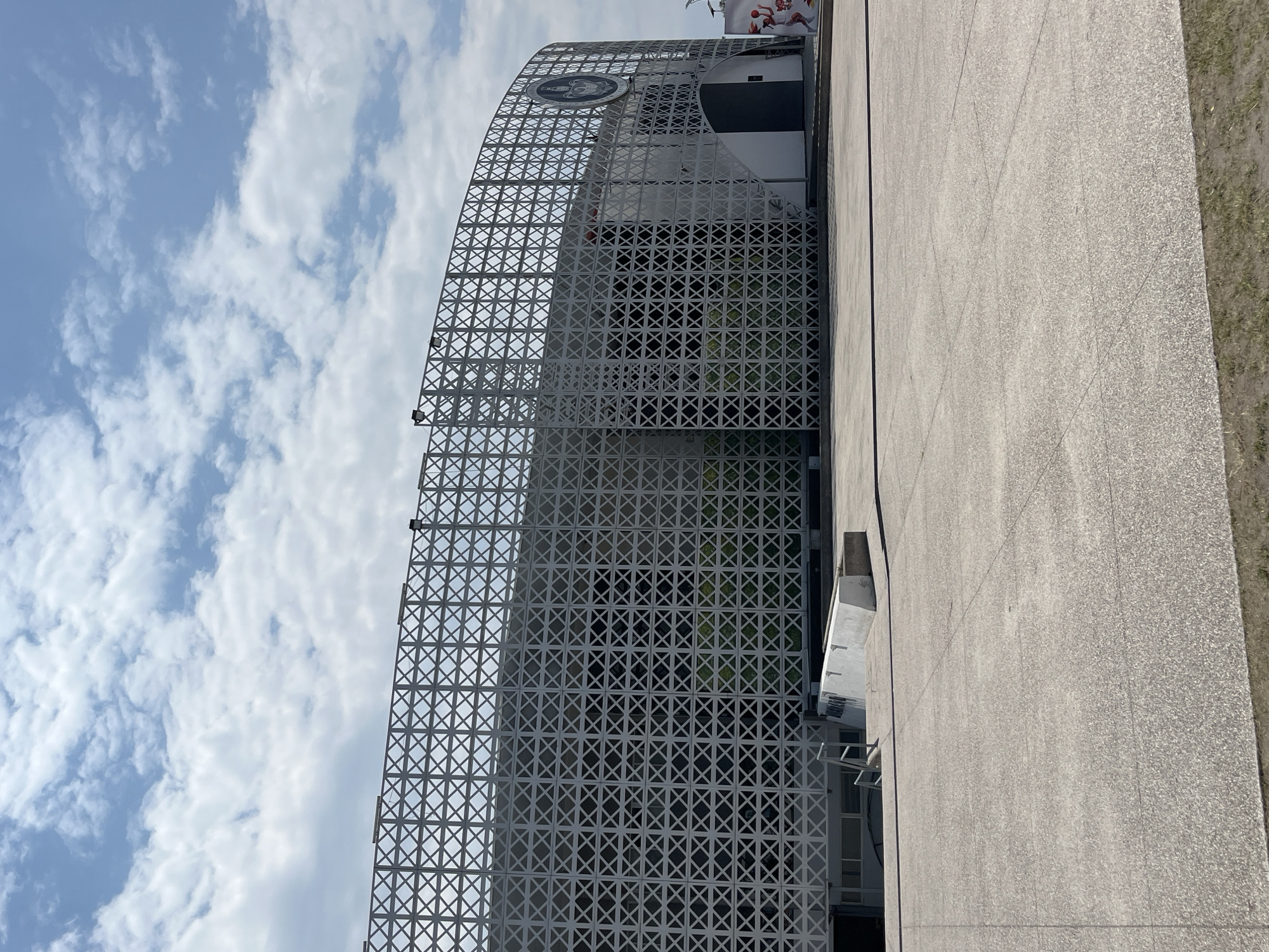 The image depicts the Frost Bank Center in San Antonio, Texas. The exterior is characterized by a distinctive curved facade covered with a white, latticework-like grid. This architectural detail extends over the building's structure. Above the latticework is an emblem. The building is adjacent to a wide concrete walkway or plaza. The sky is partly cloudy with scattered white clouds against a light blue background.