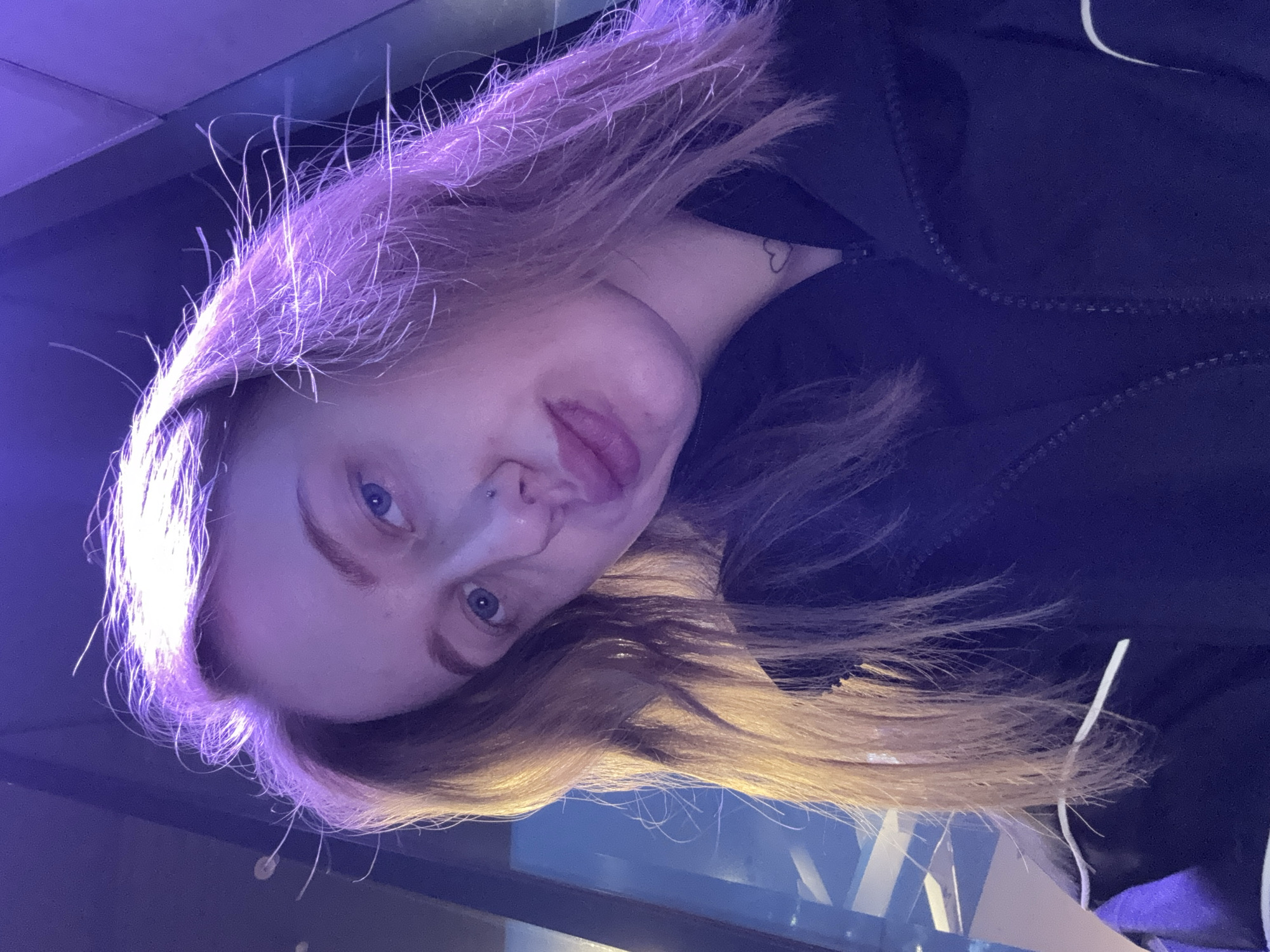 A portrait of a fair-skinned person with light blue eyes and light brown hair under violet-toned lighting. The person has a nose piercing on the left side and a heart tattoo on their neck. They are wearing a dark, possibly black, zippered jacket. The background appears to be a combination of dark and light blue areas, possibly indicating an indoor setting with some reflective surfaces. The image is taken from a low angle, looking upwards towards the person. The city and location are not discernible from the image.