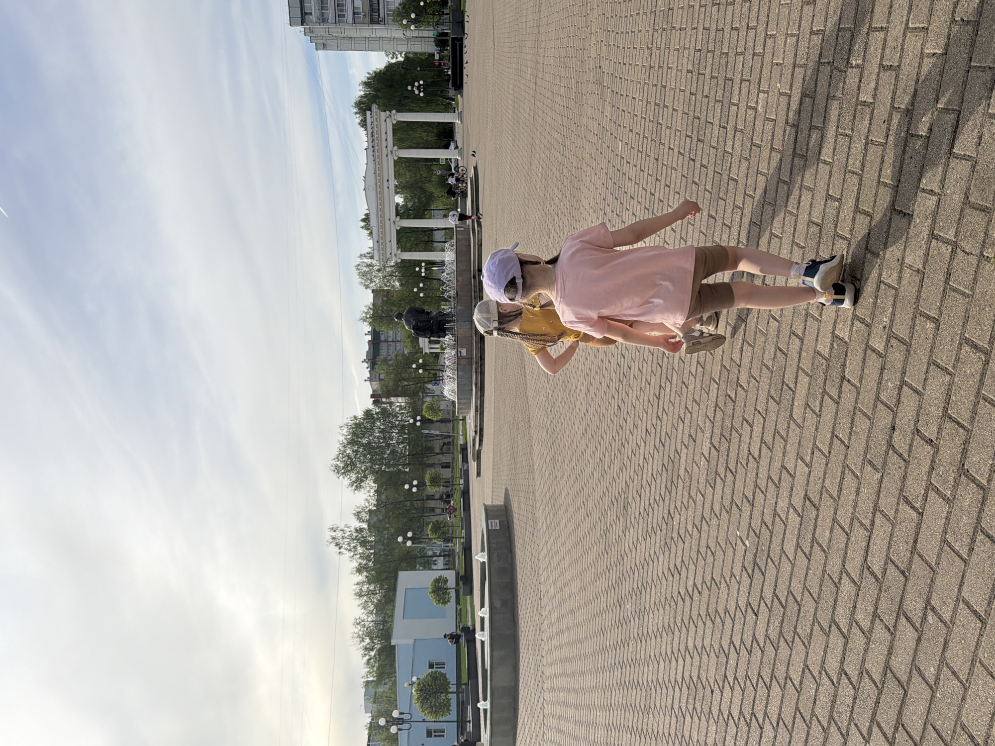 Two young children stand on a paved square in Chaikovsky, Russia. The older child, wearing a pink shirt, khaki shorts, and a lavender cap, extends their arms. The younger child, wearing a yellow shirt, khaki shorts, and a light-colored cap, also extends their arms. The square is paved with rectangular stones. In the background, there are trees, a fountain, white columns, and buildings. The sky is partly cloudy.