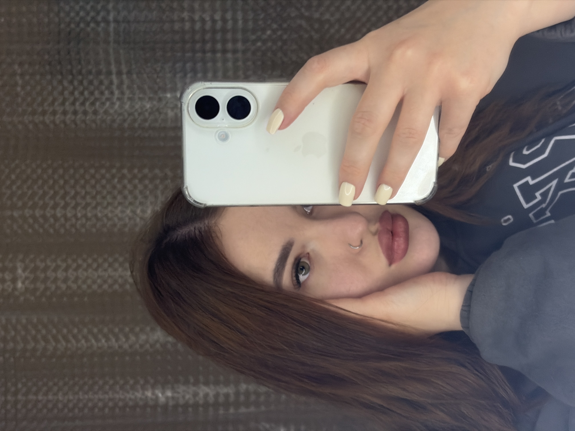 A woman is taking a selfie with an iPhone. The phone is white with a clear case and is held directly in front of her face, partially obscuring it. Her left eye is visible, as well as her nose, lips, and part of her forehead. She has a silver nose ring. Her hair is brown and falls around her face. Her fingernails are painted a light, neutral color. She is wearing a dark-colored sweatshirt with white writing. The background appears to be a textured wall or surface. The location in the city of Krasnodar, Russia, cannot be determined from this image.