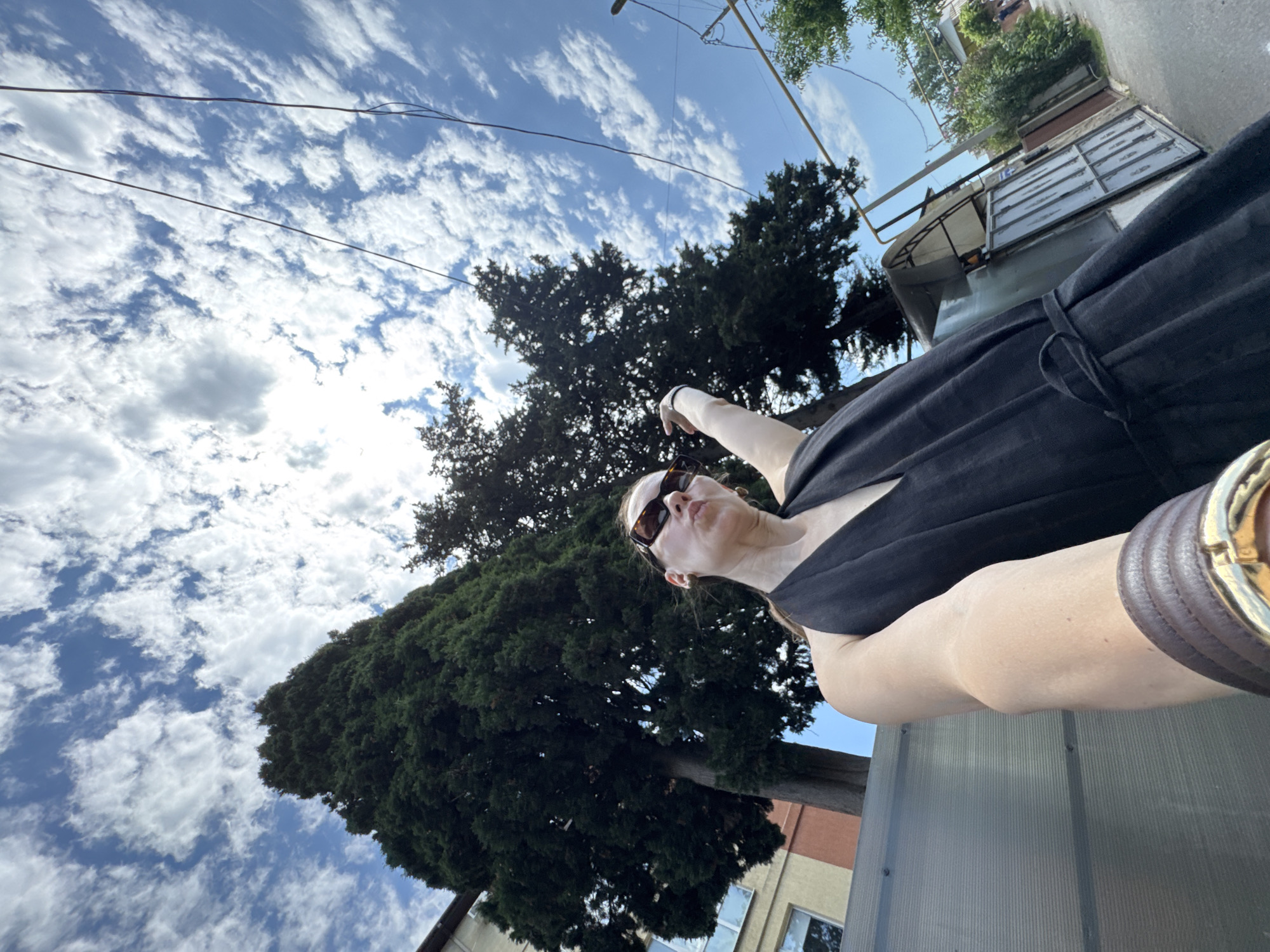 In a selfie taken from a low angle in Sochi, Russia, a woman wearing a black halter dress and sunglasses poses under a bright blue sky with scattered white clouds. She is standing in front of a tall, dark green tree and a building. Her left arm is extended, and her hand is pointing upwards, with her fingers splayed. She is looking towards the camera with her lips pursed. A wide, brown leather bracelet with a gold clasp adorns her wrist. The background shows a street with a fence and more trees.
