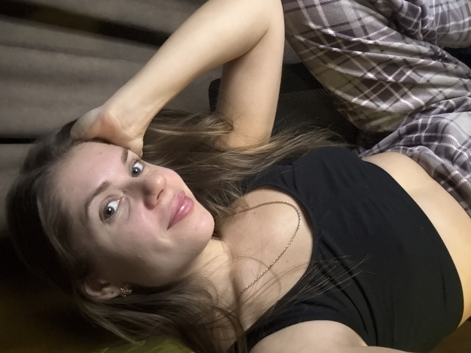 A woman lies at an angle, facing the camera with a slight smile. She wears a black tank top and plaid pajamas. Her left arm is raised above her head, hand resting on the surface behind her. A delicate gold necklace adorns her neck. Her hair is long and light brown. An earring is visible in her left ear. The background is blurred and indistinct.
