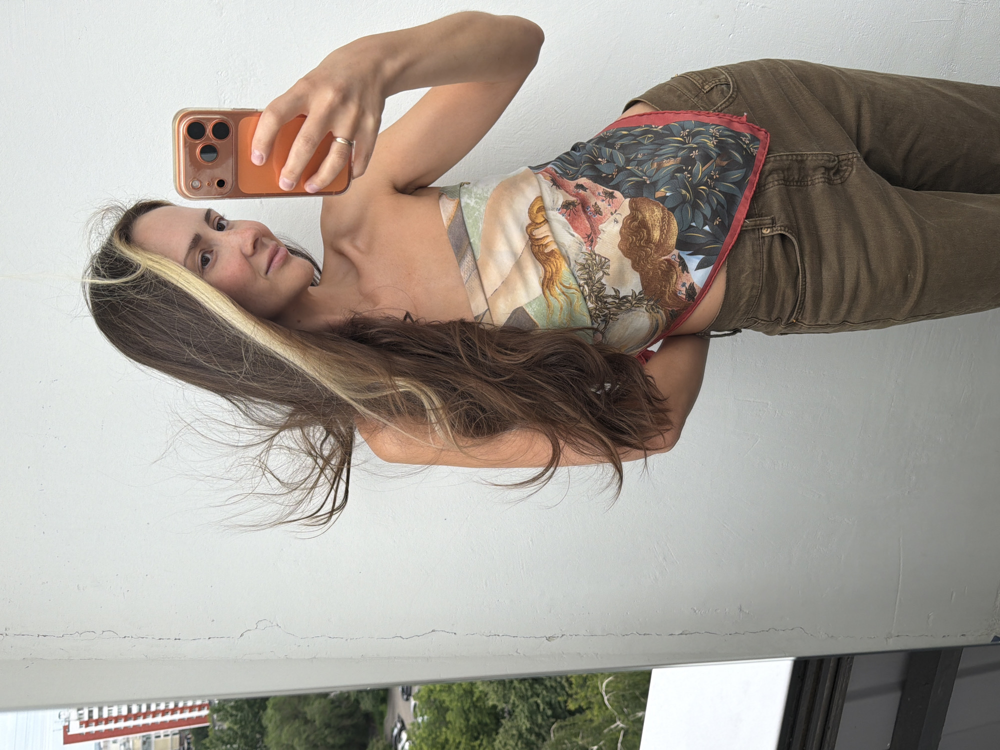 This media file is a portrait of a young woman taking a selfie with her phone. She is wearing a patterned silk scarf as a top and brown jeans. The scarf has a Renaissance-style print, featuring figures and foliage. Her hair is long and dark brown with blonde highlights, and it is flowing. She has a ring on her finger.
The woman is standing against a plain white wall, which appears to be indoors or on a balcony. In the background, visible through an opening, is a view of trees and buildings. The buildings suggest an urban or suburban setting, and the presence of trees indicates it is likely daytime and possibly spring or summer, given the foliage. The lighting is natural, suggesting daylight. The woman's expression is relaxed and she appears to be posing for the photo.
The media file is oriented vertically, indicating it was likely taken with a phone in portrait mode. The image is a close-up, focusing on the woman from the waist up, with her arm extended to hold the phone. There is no visible text in the image. The setting suggests a casual, everyday moment.