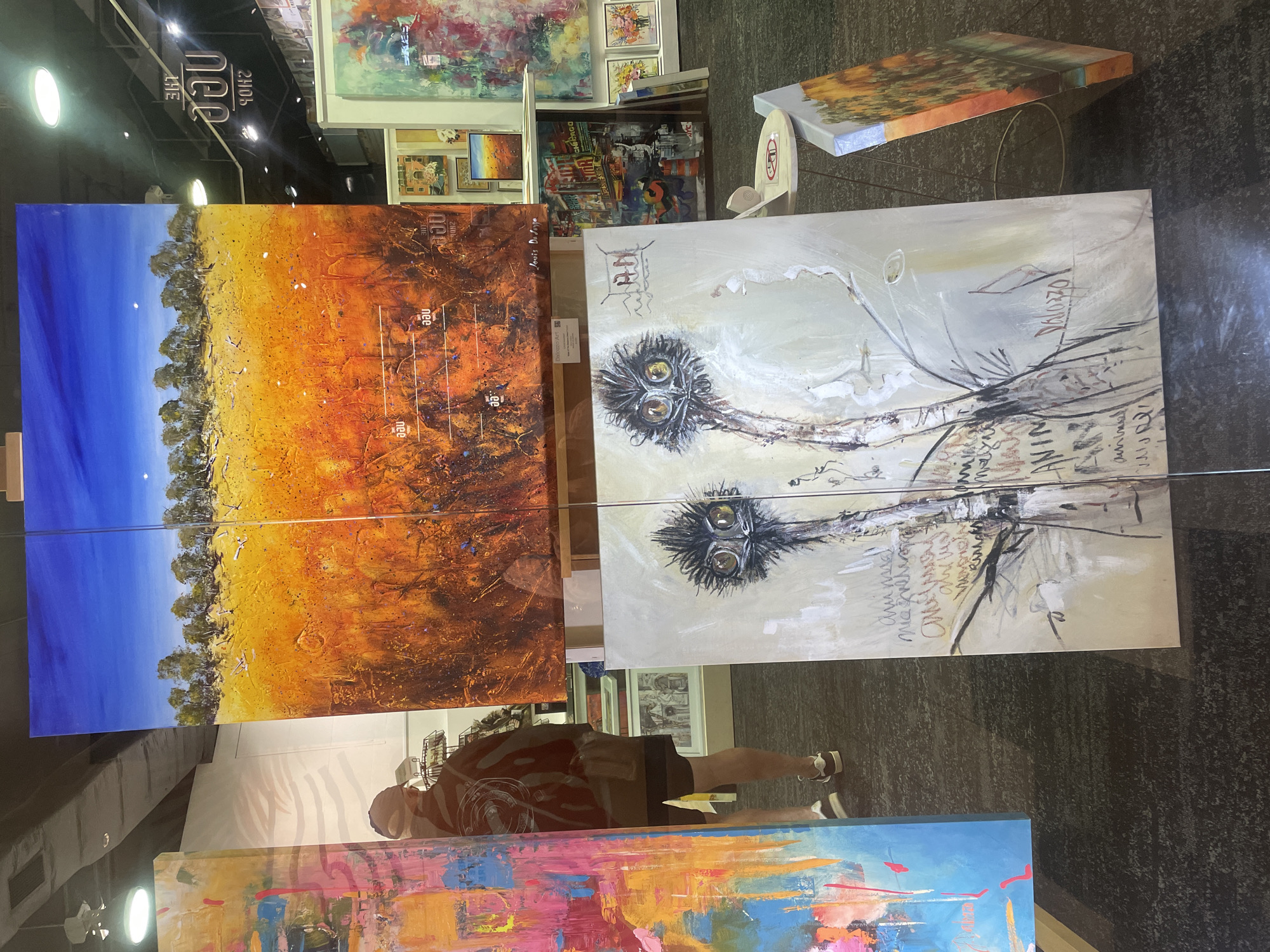 The image shows an art exhibition at the "Dialogue Art" gallery in Surfers Paradise, Australia. In the foreground, a large painting of an emu with distinctive yellow eyes is displayed. To the left, another abstract painting with vibrant colors is visible. Behind these, a panoramic landscape painting of an Australian outback scene with a bright blue sky dominates the upper portion of the image. In the background, more artwork and a person browsing are visible. The exhibition appears to be in a well-lit indoor space with a dark carpeted floor.