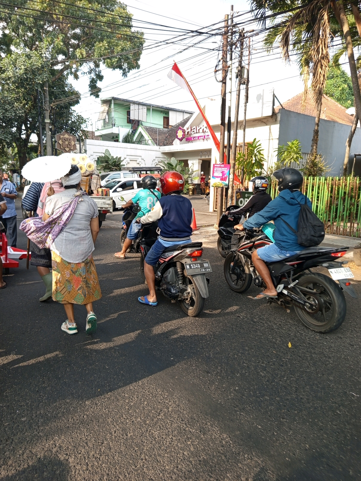 The image depicts a street scene in Malang, Indonesia. In the foreground, a woman wearing a batik skirt and a white headscarf is walking away from the camera, carrying a white umbrella. She has a patterned cloth slung over her shoulder. To her left, another woman, partially obscured, also carries a white umbrella. Behind them, several motorcycles are visible. One motorcycle, ridden by a person in a turquoise shirt with the number "88", is in the middle ground. To the right, another motorcycle with a rider wearing a blue jacket and a black backpack is also in motion. In the background, buildings with red-tiled roofs and a white facade with a sign that reads "Haris Cafe and Resto" are visible. Power lines crisscross the sky, and lush green trees line the street. The overall lighting suggests it is daytime, with strong shadows cast on the asphalt road.