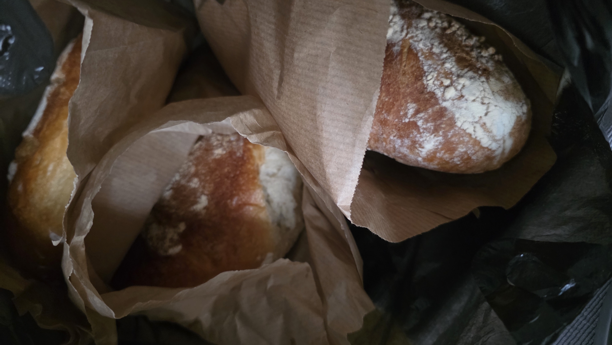 The image shows three loaves of bread in paper bags inside a black plastic bag. The bread loaves are crusty and appear to be dusted with flour. The paper bags are crumpled and slightly open, revealing the bread inside. The black plastic bag is shiny and partially obscures the contents. The setting appears to be indoors, based on the lighting and lack of identifiable outdoor elements. There is no indication of the location being in Blagoevgrad, Bulgaria. No people are visible in the image.