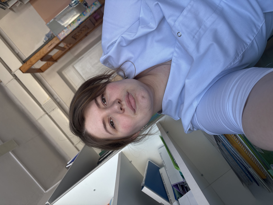 A woman with brown hair and fair skin wears a white coat or uniform, possibly in a medical or academic setting. She is positioned in front of white bookshelves filled with books and folders. The photo's angle suggests she's holding the camera, creating a selfie-style shot. The background also shows a high wooden structure.