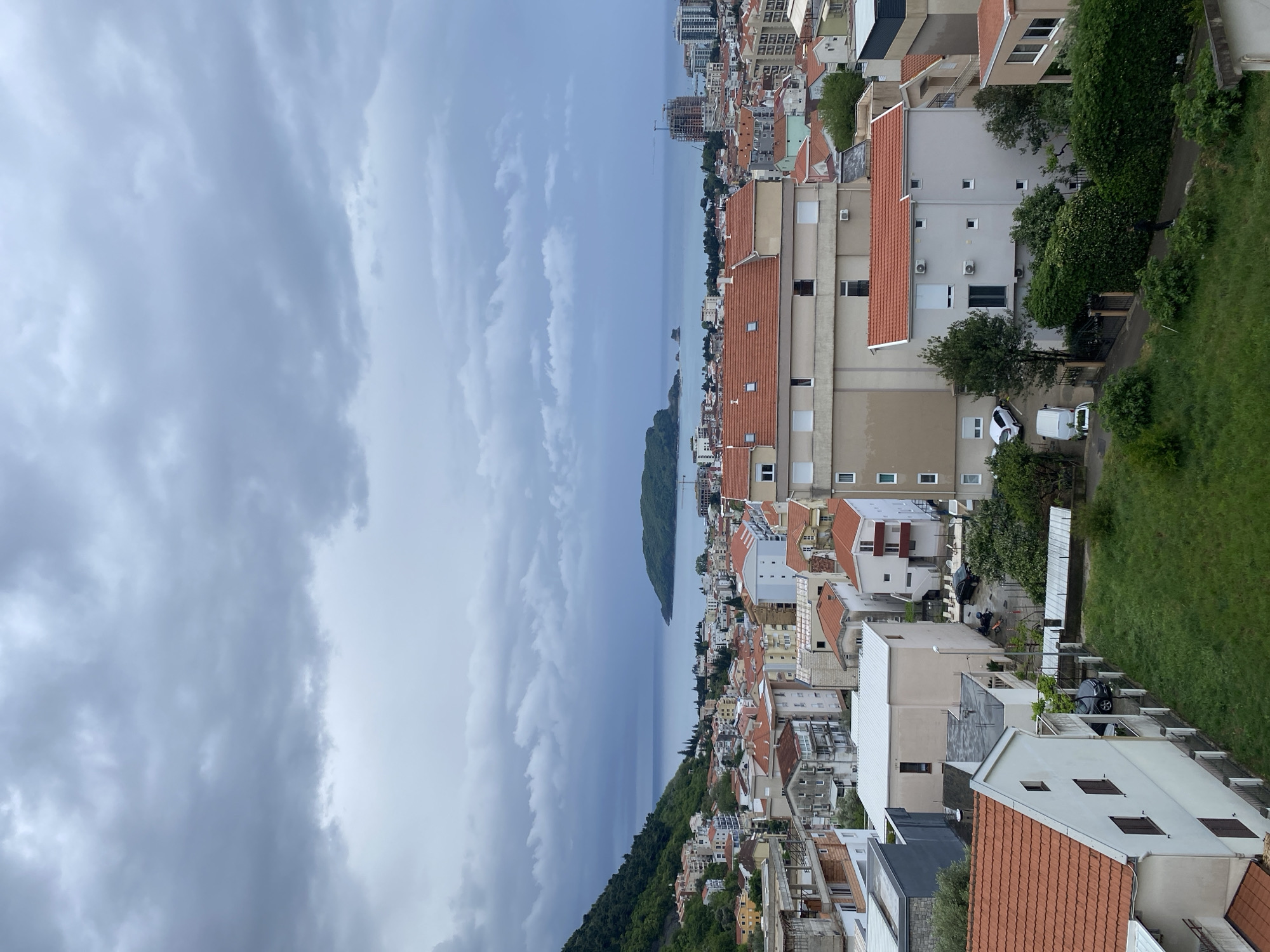 The image presents an elevated, panoramic view of a densely populated coastal town, likely Budva, Montenegro, under an overcast sky. The scene is dominated by a calm, grey-blue body of water, possibly the Adriatic Sea, stretching to the horizon. A small, verdant island or landmass is visible in the distance, centered in the water.
The town itself is characterized by numerous buildings, mostly residential, featuring distinctive reddish-orange tiled roofs. The structures exhibit varied facades, predominantly in shades of white, beige, and light grey. Many are multi-story, some with visible windows and balconies. Towards the background, taller, more contemporary buildings and visible construction suggest ongoing urban development.
Green vegetation is abundant, with trees, bushes, and grassy slopes interspersed among the buildings and covering the hillsides that frame the town. Streets and pathways are discernible, with several parked vehicles, including a white van and a black car, hinting at daily life. A few small, indistinct human figures are faintly visible in some areas, but no significant activity is taking place.
The sky is heavily clouded with dense, grey formations, indicating overcast or possibly pre-rainy weather. The diffused lighting suggests it is daytime, though lacking direct sunlight. The overall atmosphere is calm and subdued due to the muted light. No discernible text is visible within the image.