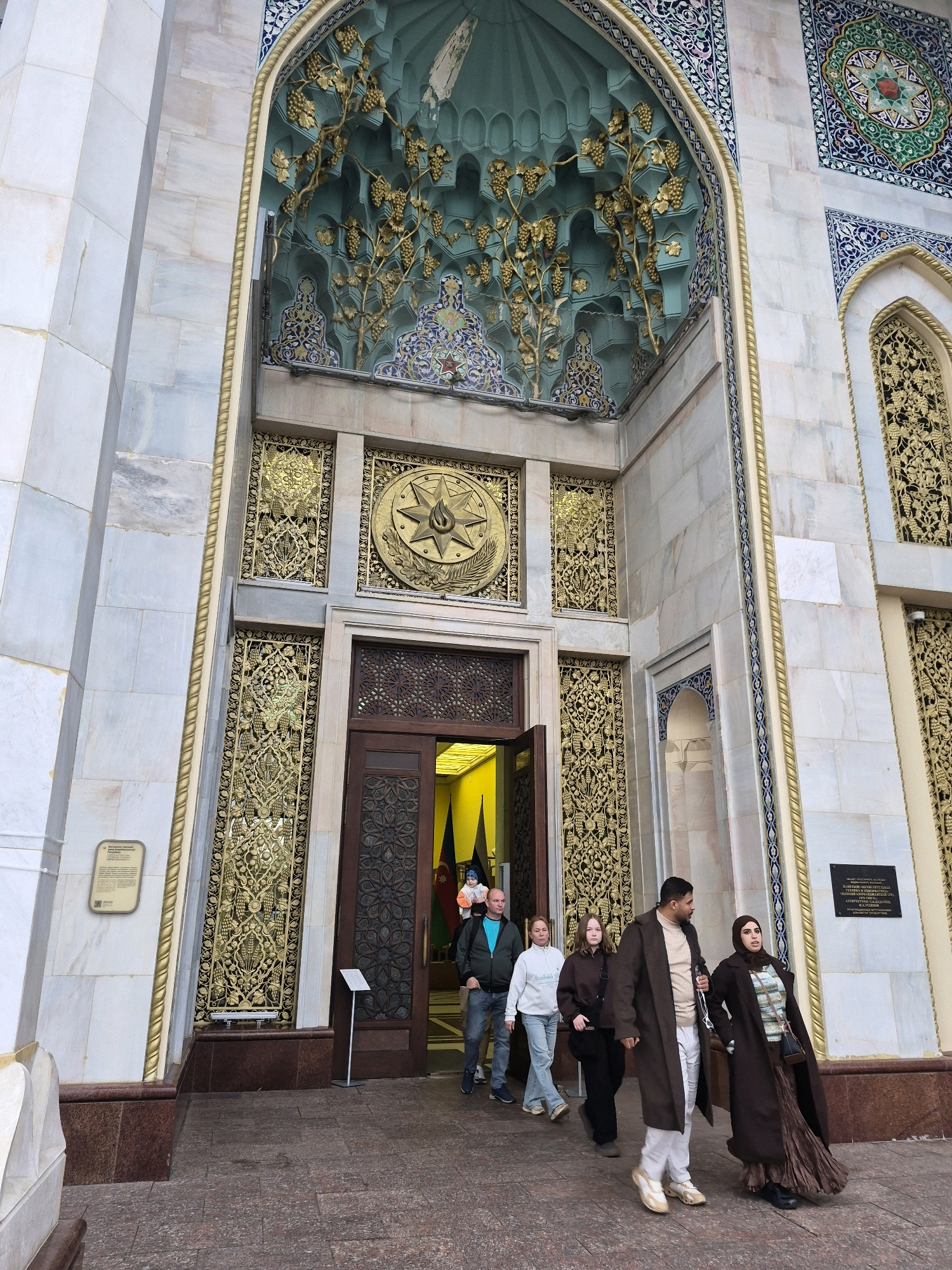 The image shows the entrance to the Azerbaijan Cultural Center in Moscow, Russia. The building features ornate architecture with marble cladding, intricate gold detailing, and mosaic work. In the foreground, a group of people is walking towards the entrance. The group consists of four individuals: a man in a dark brown coat and white pants, a woman in a dark brown coat and a long brown skirt, a young woman in a light-colored hoodie and jeans, and another young woman in a dark hoodie and black pants. They are walking down a stone pathway leading to the building. The entrance itself is recessed and features a large wooden door with intricate carvings. The overall scene depicts a cultural landmark with visitors entering the building.