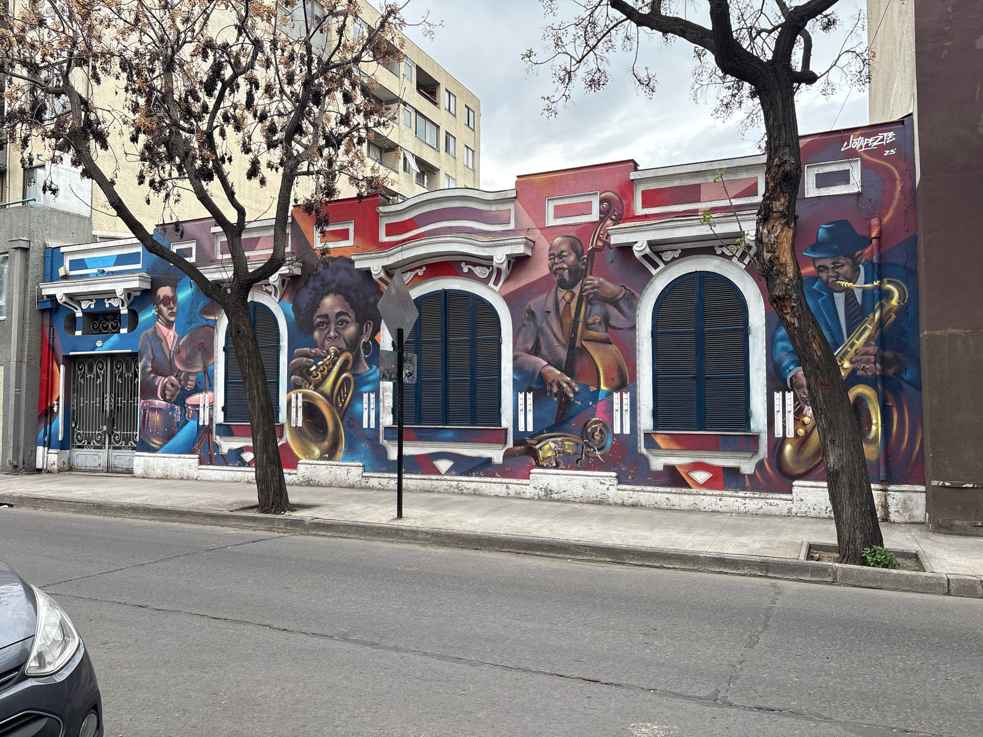 This is a street-level, medium shot of a mural on the side of a building in Santiago, Chile. The mural depicts several jazz musicians playing instruments. On the left, a drummer is shown with his hands over his drums. In the center, a woman with an afro plays a saxophone. To the right, a man plays a double bass, and another man in a blue suit and hat plays a saxophone. The mural is vibrant and colorful, with red and blue as dominant colors. The building itself appears to be a multi-story structure with balconies. There are trees in the foreground partially obscuring the mural. A gray car is visible in the lower left corner of the image. The street in front of the building is asphalt, and there is a sidewalk. The sky is overcast. The mural includes the artist's signature, "LOTAPEPE 23".