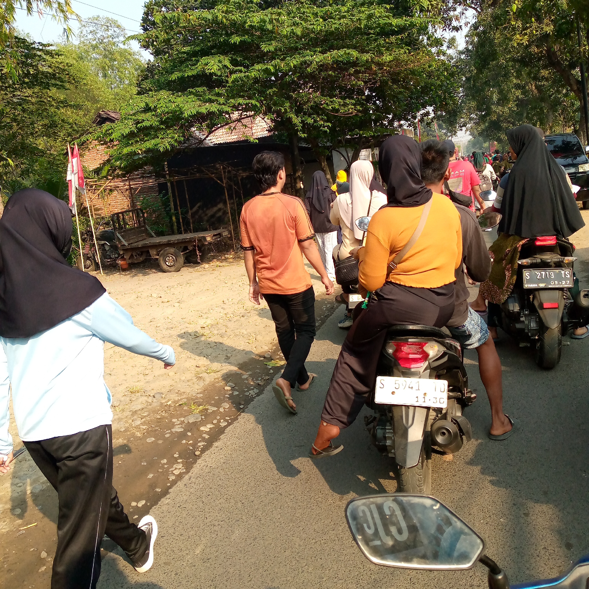 A group of people are on a paved road under a canopy of trees with a building with a red tile roof in the background. The scene appears to be in Tambakagung, Indonesia. One person in the foreground, facing away from the camera, is wearing a light blue long-sleeved shirt and black pants with white stripes, and white sneakers. To the right of this person, two individuals are on a motorcycle; the person in front is wearing a black headscarf and an orange long-sleeved shirt, and the person behind them is wearing a black headscarf and a green patterned garment. To the right of them, another motorcycle has a rider in a black headscarf and an orange top, and the passenger behind them is wearing a black headscarf and a patterned green garment. A black SUV is visible further down the road on the right. A wooden cart with one wheel is parked on the side of the road to the left of the frame, near a brick building with a thatched roof. The road is dirt and gravel on the sides, with a paved surface in the middle. The sky is clear and bright, suggesting it is daytime.