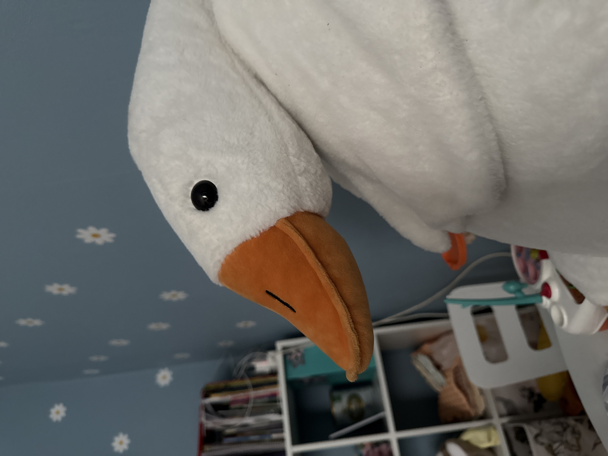 A close-up, upward-angled shot features a white, plush goose toy. The goose has an orange beak with a black line detail and a small black eye. The background reveals a light blue wall decorated with white daisy patterns, and a white shelving unit filled with various objects. The toys are behind the toy goose. There is no indication of Moscow or Russia in this image.