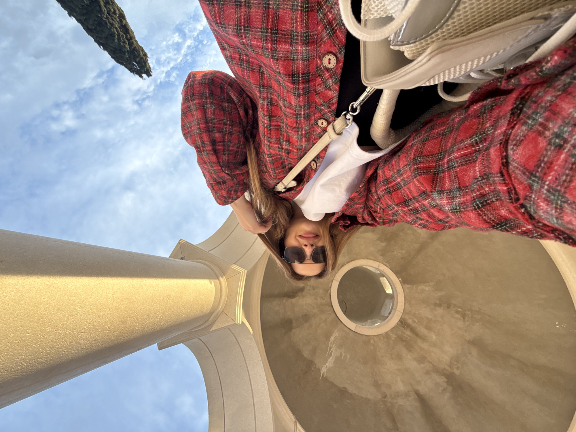 A low-angle shot presents a woman in sunglasses, wearing a red plaid shirt over a black top and white undershirt. She’s standing beneath an architectural structure in Đuraševići, Montenegro, featuring a colonnade and a domed ceiling with a circular window. The background is a blue sky with wisps of clouds, and a tree branch is visible at the upper left. The architecture is light in color, possibly stone or concrete, and sunlight creates shadows on the surfaces.
