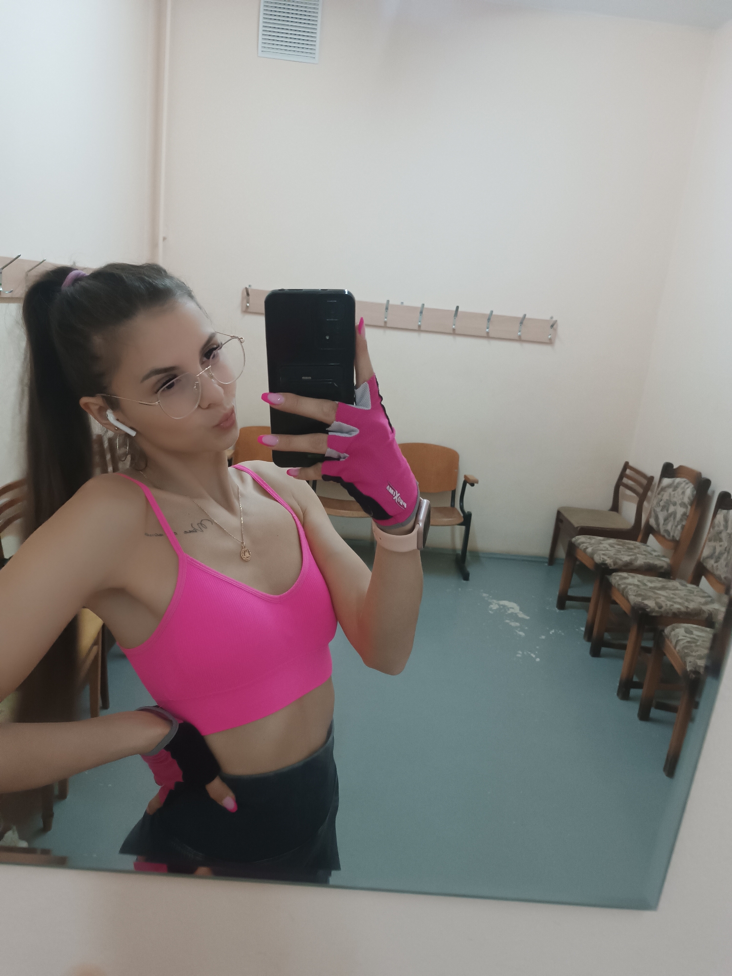 A young woman with a high ponytail and eyeglasses is taking a mirror selfie in a room that appears to be a changing room or a small waiting area. She is wearing a bright pink sports bra, black shorts, and pink fingerless workout gloves. She has on white wireless earbuds and a delicate gold necklace. The room has pale walls with a row of hooks on the upper wall. In the reflection, several wooden chairs with patterned cushions are visible, as well as a light blue floor.