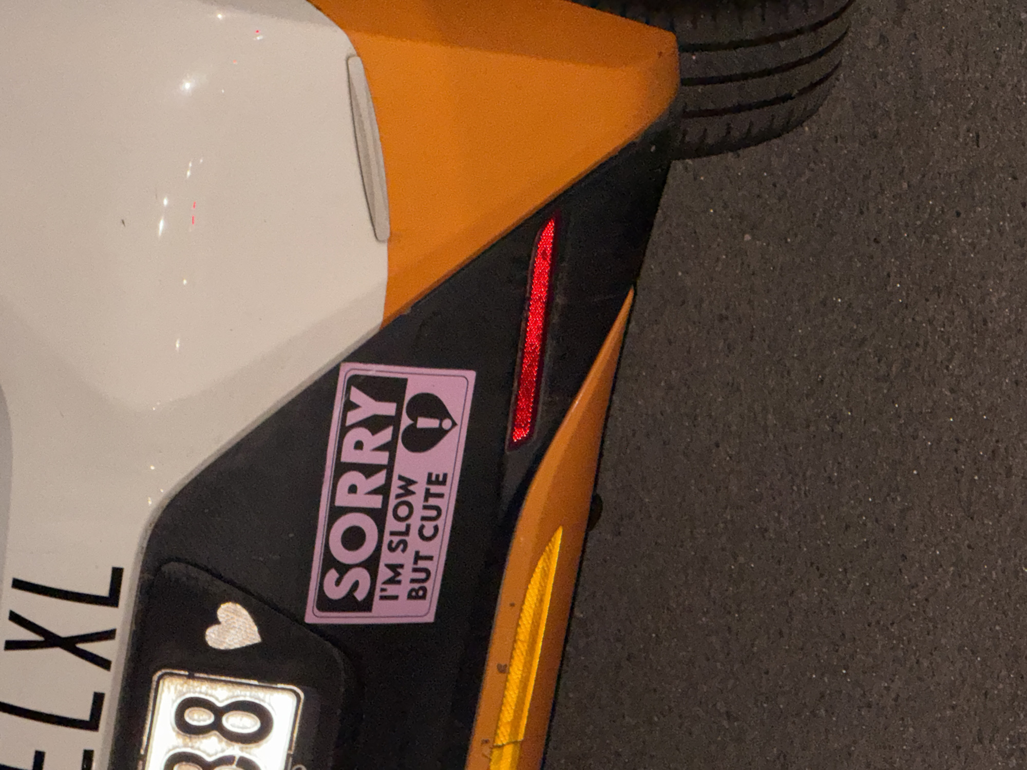 The image depicts a portion of a vehicle, likely a car, parked on a paved surface. The visible section of the car is located near the front driver's side wheel well. The car has a white and orange paint scheme with a black accent. A pink sticker is affixed to the black section, reading "SORRY I'M SLOW BUT CUTE" with a heart and question mark graphic. A silver heart-shaped sticker is visible near the license plate, which partially reads "XL 8". A small vertical red reflector is also present. The tire of the vehicle is partially visible at the top of the frame, indicating the car is parked on a road or parking space. The location is given as Borough of Spelthorne, United Kingdom but there are no aspects of the image which indicate this is the exact location.