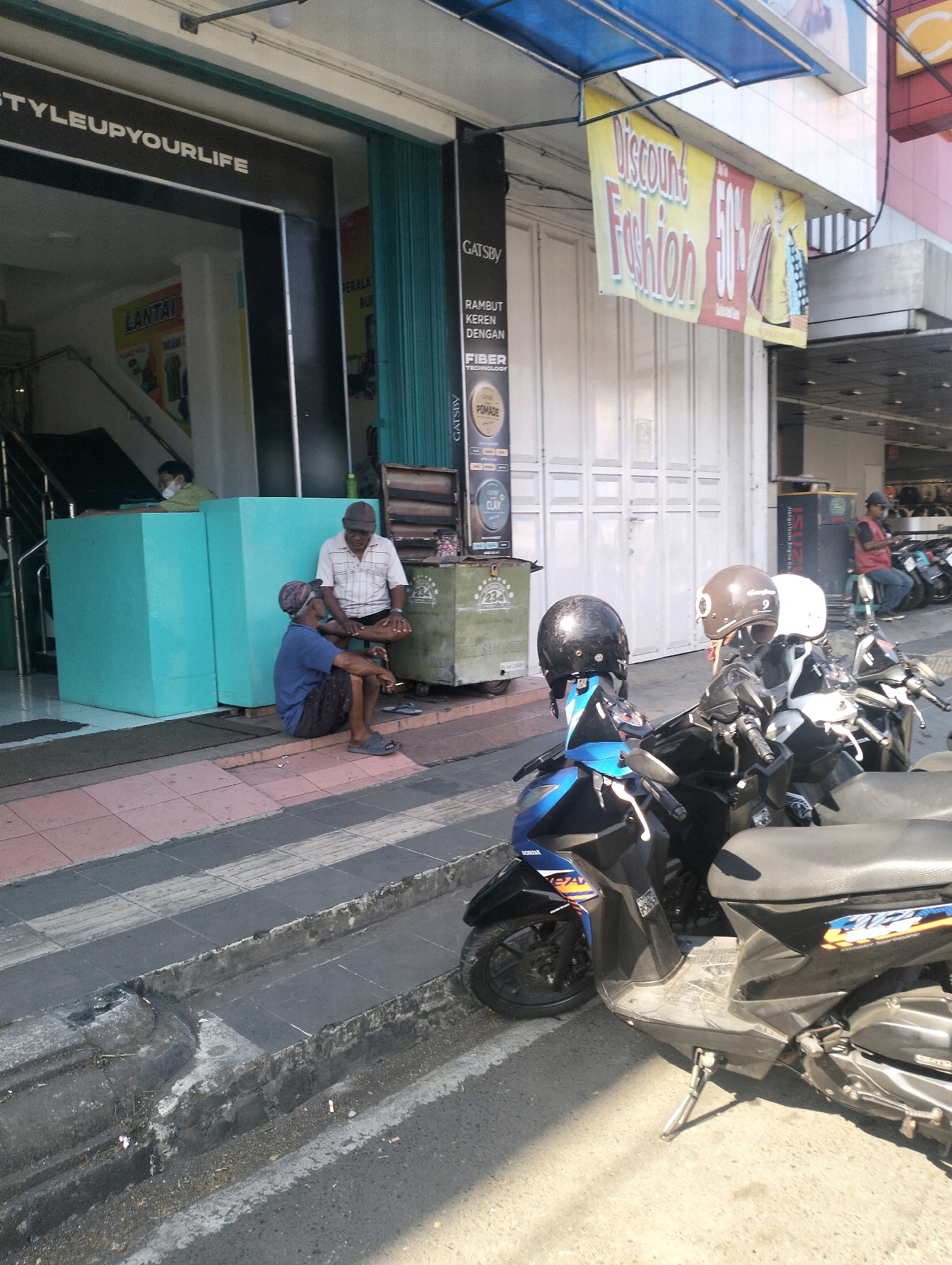 The image captures a street scene in front of a store in Purbalingga, Indonesia. The store's facade features a large sign with the text "STYLEUPYOURLIFE." To the right of the entrance, a yellow banner advertises "Discount Fashion 50%."
Two men are seated on the sidewalk in front of the store. One man, wearing a blue shirt and patterned shorts, sits on the ground. The other man, in a white and striped short-sleeved shirt and dark shorts, is seated on a low stool or box. They appear to be engaged in conversation.
Several motorcycles are parked on the street in the foreground, some with helmets placed on them. A person wearing a red vest is visible further down the street, near another motorcycle.
The environment includes a sidewalk with a patterned tile section, a concrete drainage channel, and an asphalt road. The buildings have white doors and a blue awning above the discount banner. The lighting suggests it is daytime with direct sunlight casting shadows.