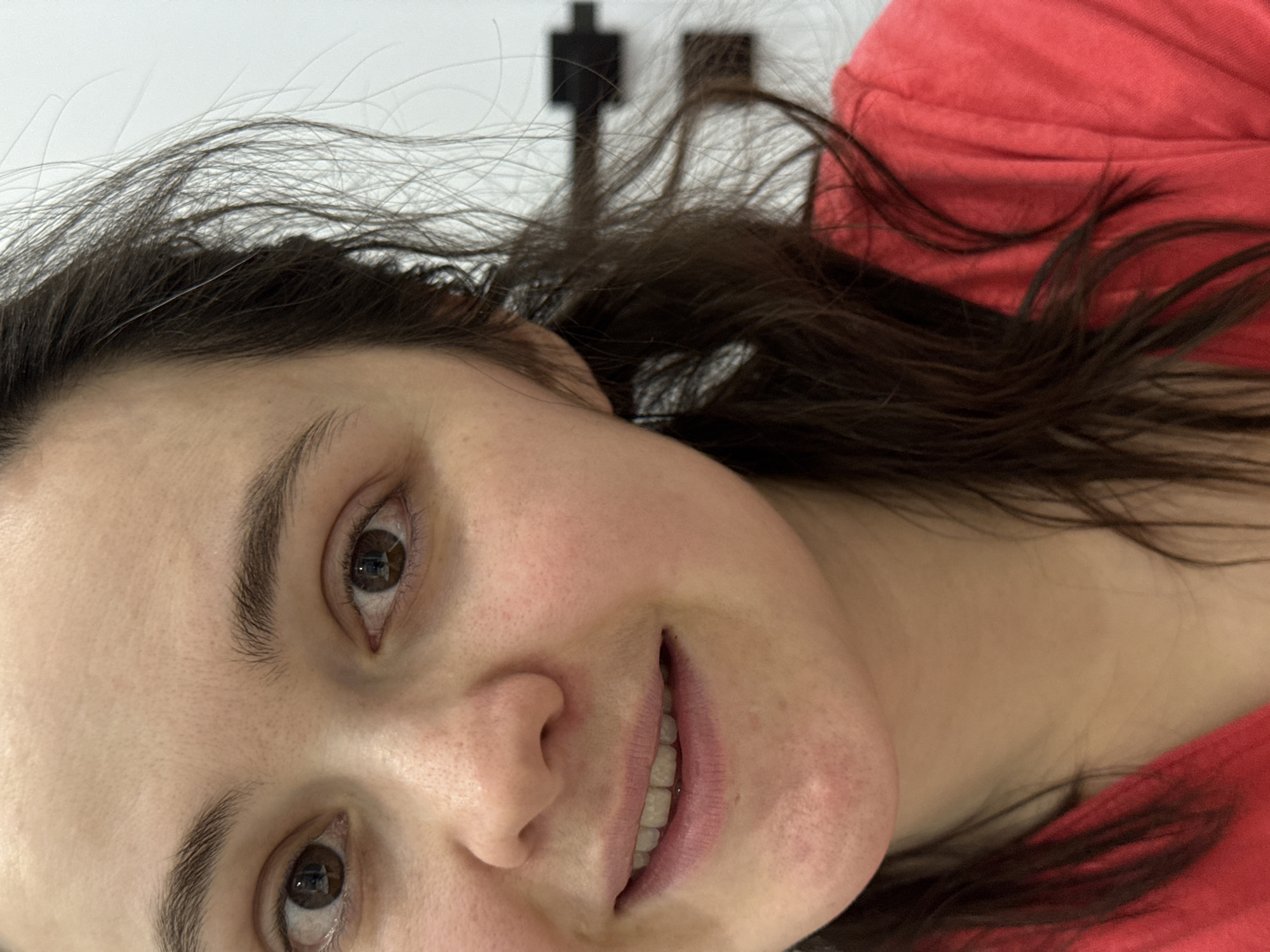 A close-up, angled portrait of a woman with fair skin, brown hair, and brown eyes. She is smiling slightly, revealing her teeth. She is wearing a red shirt. The background is blurred and includes a white wall and a black object attached to the wall. There is no information about this image being in the city of Izhevsk, Russia.