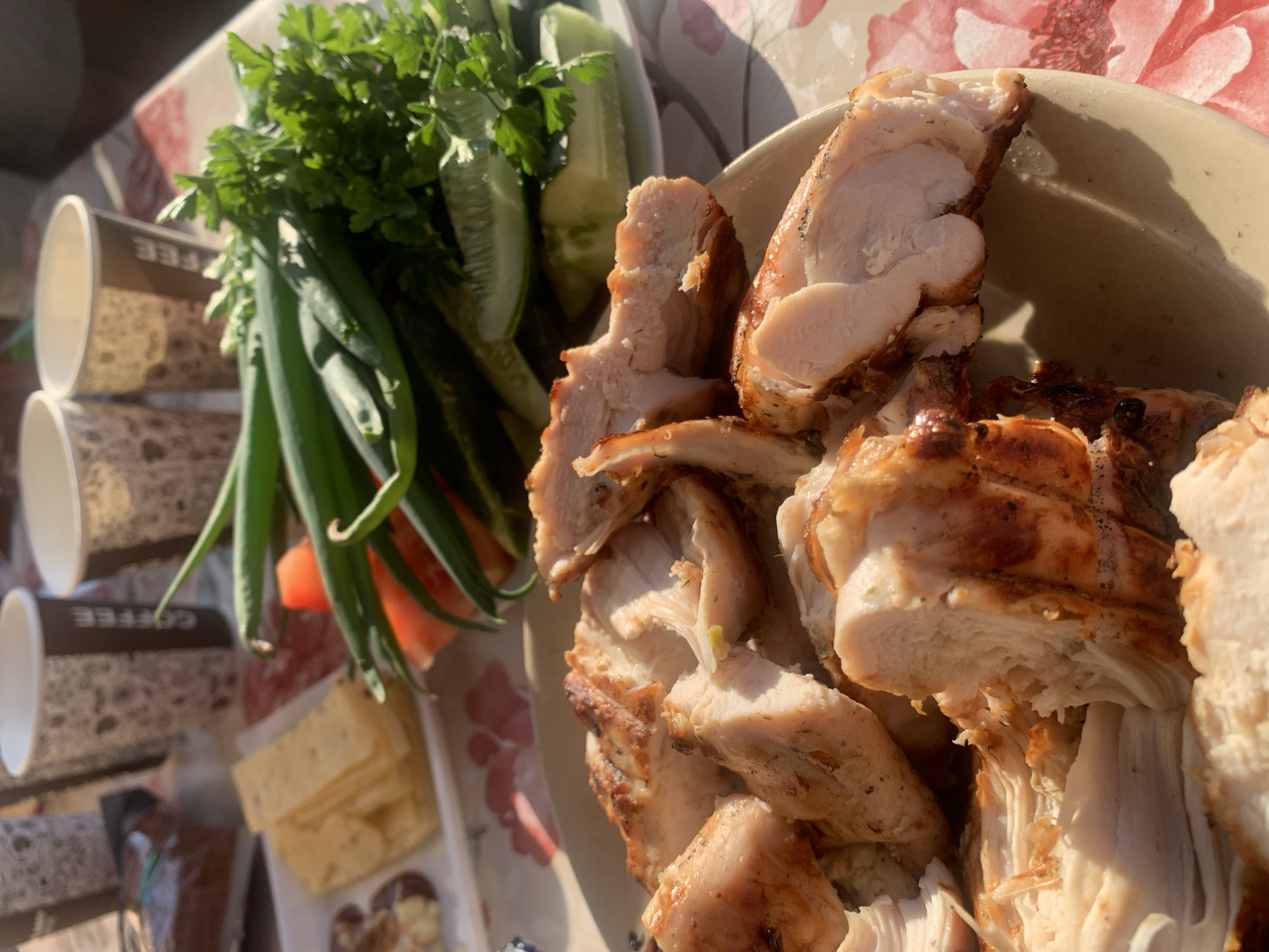 This image shows a plate of sliced grilled chicken next to a collection of fresh vegetables, including green onions, cucumbers, and parsley. There are also several disposable coffee cups and some slices of cheese. The scene appears to be an outdoor meal setting. There is no evidence to specify the location is in the city of Chaikovsky, Russia.