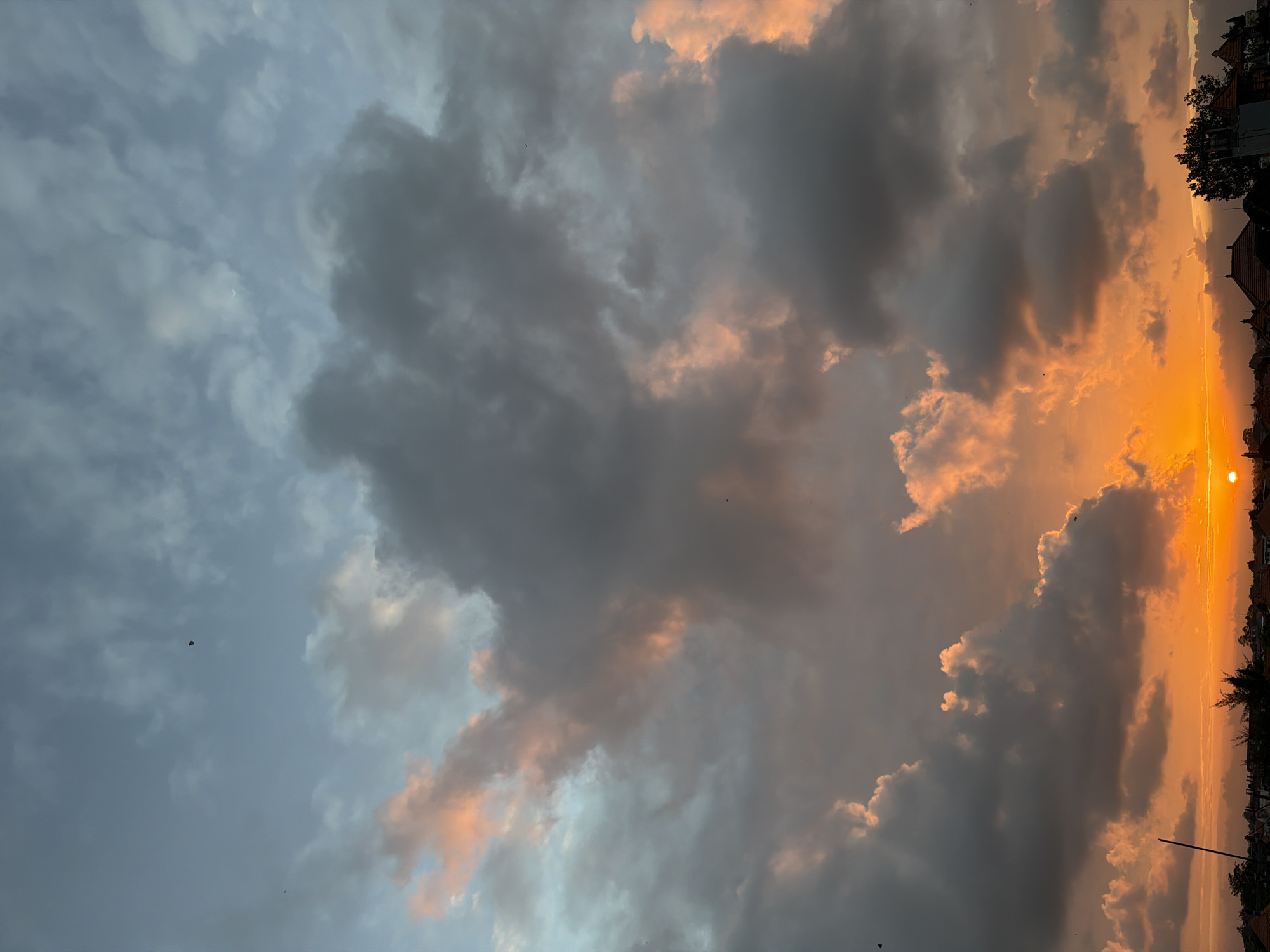 A wide shot captures a sunset in North Kuta, Indonesia, with a vibrant orange glow illuminating the horizon. The sky is filled with a dramatic mix of dark gray and light pink clouds, creating a textured and dynamic atmosphere. Silhouetted against the bright sky are rooftops of buildings, a few trees, and a thin pole reaching upwards. Several small dark specks, likely birds or insects, are scattered across the sky, adding a sense of scale to the vastness of the scene. The sun is visible as a bright orange disc peeking out from behind the clouds near the bottom right of the frame.