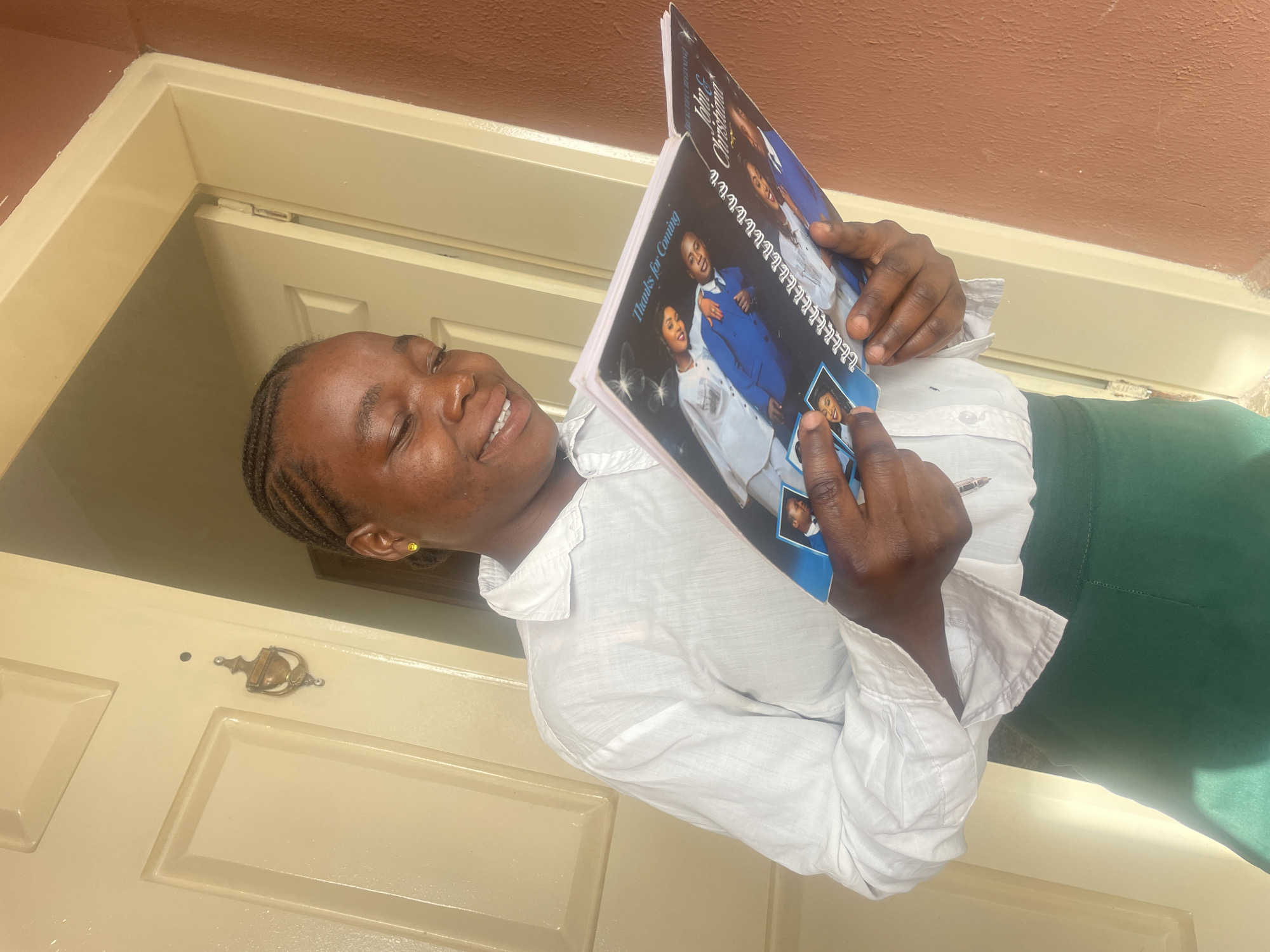 A young woman with dark skin and braided hair is depicted in a brightly lit, close-up shot, angled diagonally. She wears a white long-sleeved button-up shirt and a dark green skirt. She is holding an open, spiral-bound book or program and looking at it with a wide, happy smile. Her right index finger points to a smaller photo on one of the pages.
The book features images of people, most notably a couple: a man in a blue suit and a woman in a white dress. Visible text on the book includes "Thanks for Coming" and "John & Christiana," suggesting it is an event program, likely for a wedding or anniversary.
The background reveals a light-colored, paneled door with an ornate brass doorknocker and a reddish-brown wall or ceiling above the door frame. The strong, direct lighting indicates it is daytime, casting clear shadows. The woman has a small, yellow stud earring in her right ear. The scene appears to take place in a doorway or entrance, possibly residential, in Jos, Nigeria. The overall mood is cheerful and engaged.