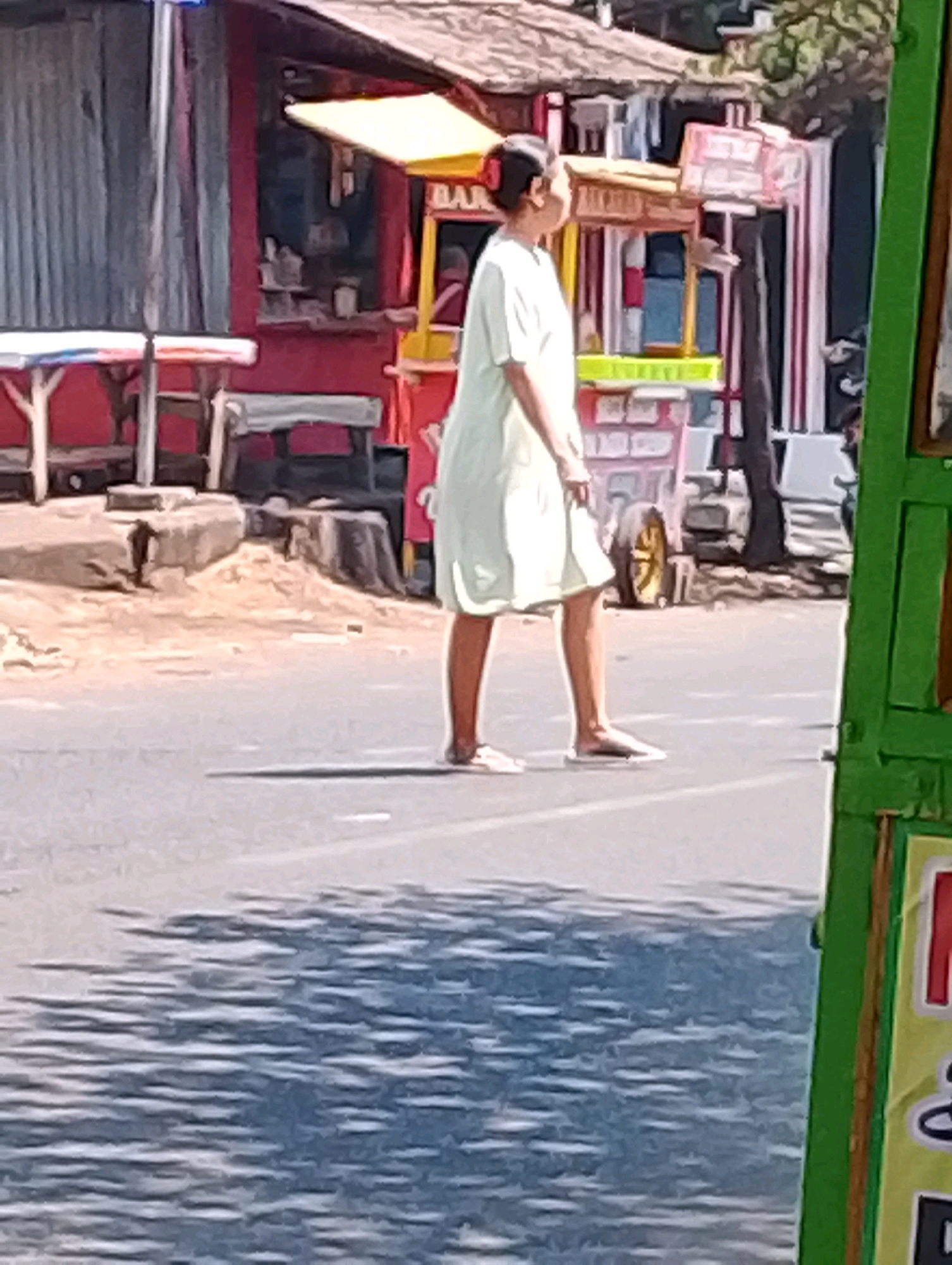 The image depicts a street scene in Probolinggo, Indonesia, likely on a sunny day given the harsh shadows. In the foreground, a person wearing a light-colored dress and flip-flops is walking on the asphalt road. The person's back is to the camera, and their hair is pulled back in a bun. The road has faint white lines indicating lanes.
In the background, there are several roadside stalls or shops. One stall, with a yellow awning, appears to be selling food or drinks. Behind it, there are other structures with red and white striped facades. The overall environment suggests a bustling street with commercial activity. The ground beside the road is rough and unpaved, with some scattered debris and large rocks. The lighting casts strong, elongated shadows from the structures and possibly trees onto the road.