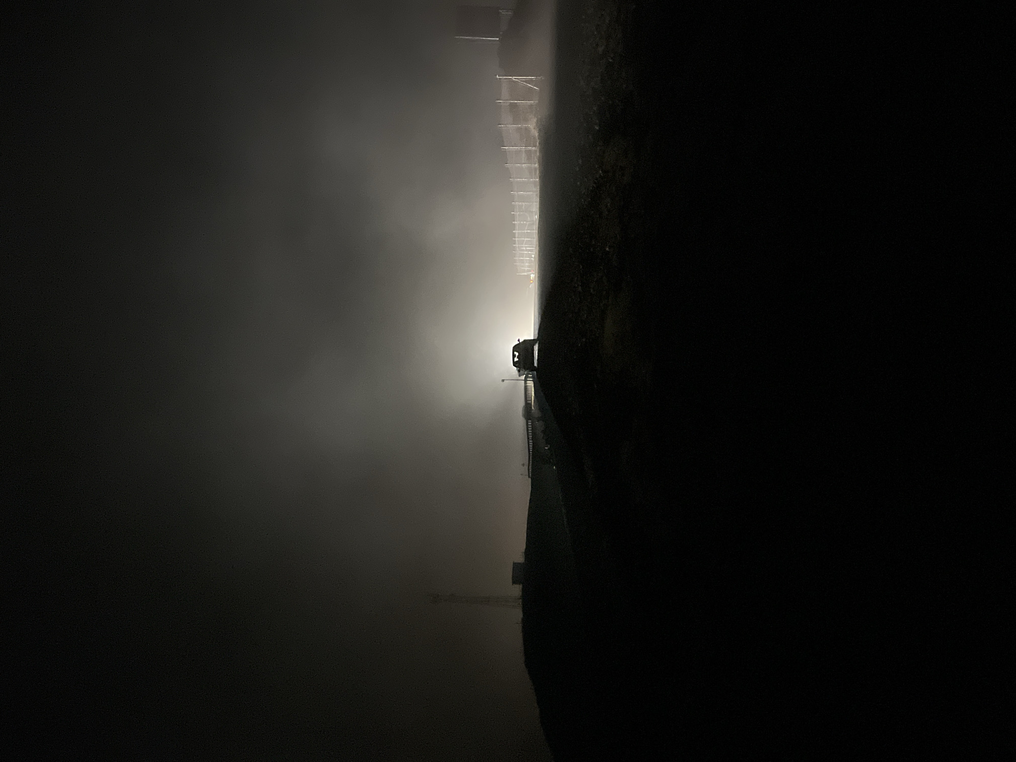 In the city of Gomismta, Georgia, a dark, foggy night scene unfolds. A vehicle, dimly illuminated by its headlights, is positioned on a road or elevated area. The fog is thick, obscuring the background and creating a hazy, ethereal atmosphere. To the left, a tall, slender structure, possibly a communication tower, is faintly visible against the dark sky. A low-lying building is situated near the base of this tower. In the mid-ground, a fence runs horizontally across the scene, its posts and wire mesh barely discernible through the mist. The ground appears to be uneven and rocky. The overall impression is one of isolation and mystery, with the fog playing a significant role in shaping the visual narrative of the scene.