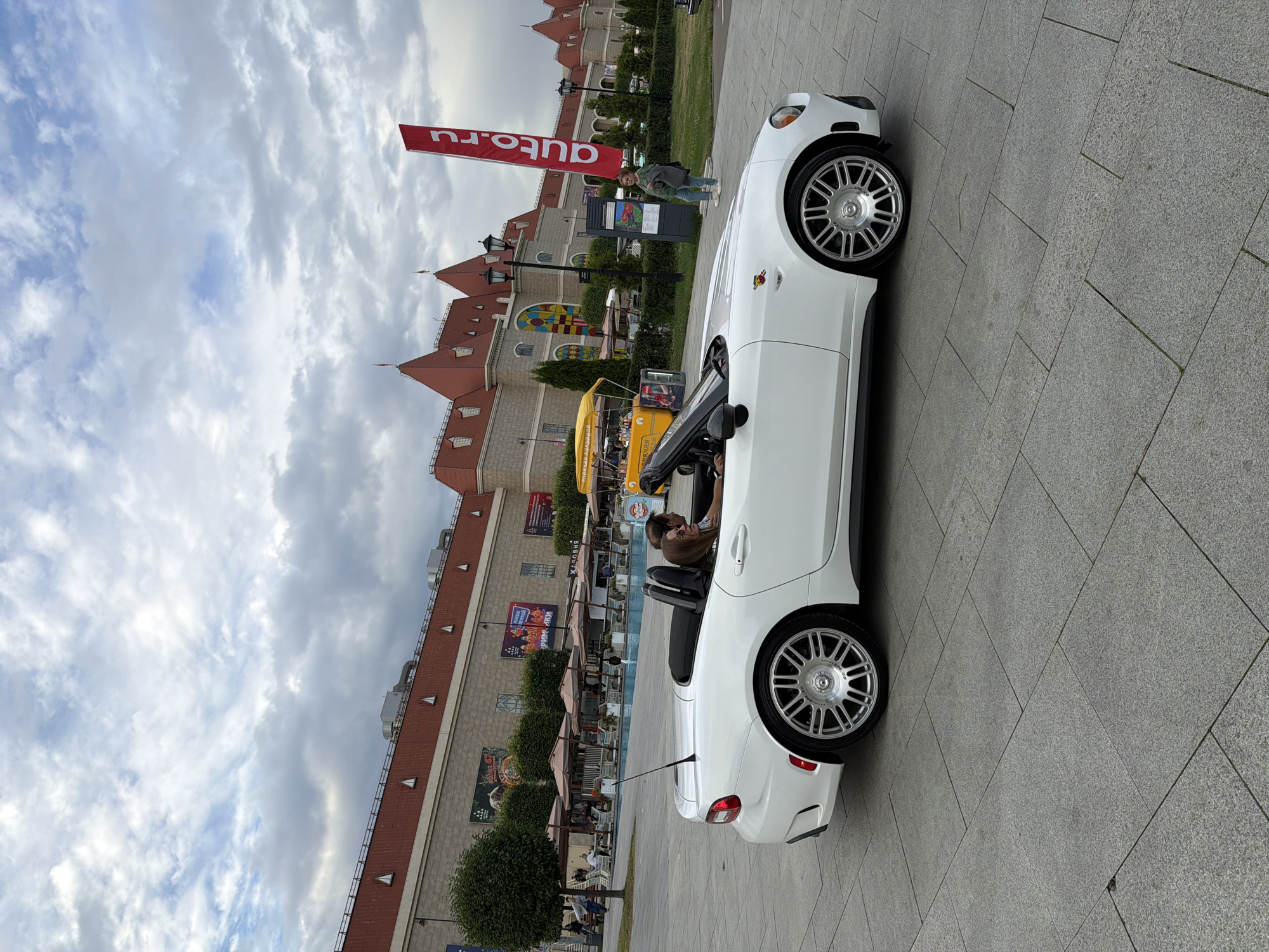 A white convertible sports car with a black top is parked on a paved plaza in front of a large building with a red roof and a beige facade. The building has several arched windows and decorative spires. A red banner with white text reading "auto.ru" is planted on the right side of the plaza. Two people are seated in the convertible, a driver and a passenger. The driver is wearing a brown shirt and the passenger is wearing glasses and a grey top. In the background, there are outdoor seating areas with umbrellas, trees, and a yellow food cart. The sky is cloudy.