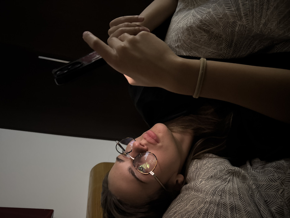 A woman with glasses lies on her back, holding a phone. Her left hand is on the phone. She wears a patterned shirt and a simple wristband. The lighting is dim, focusing on her face and hands. A dark surface looms overhead. Part of a chair is visible behind her head. The background includes a cream-colored wall. A red object is in the lower left corner of the frame.