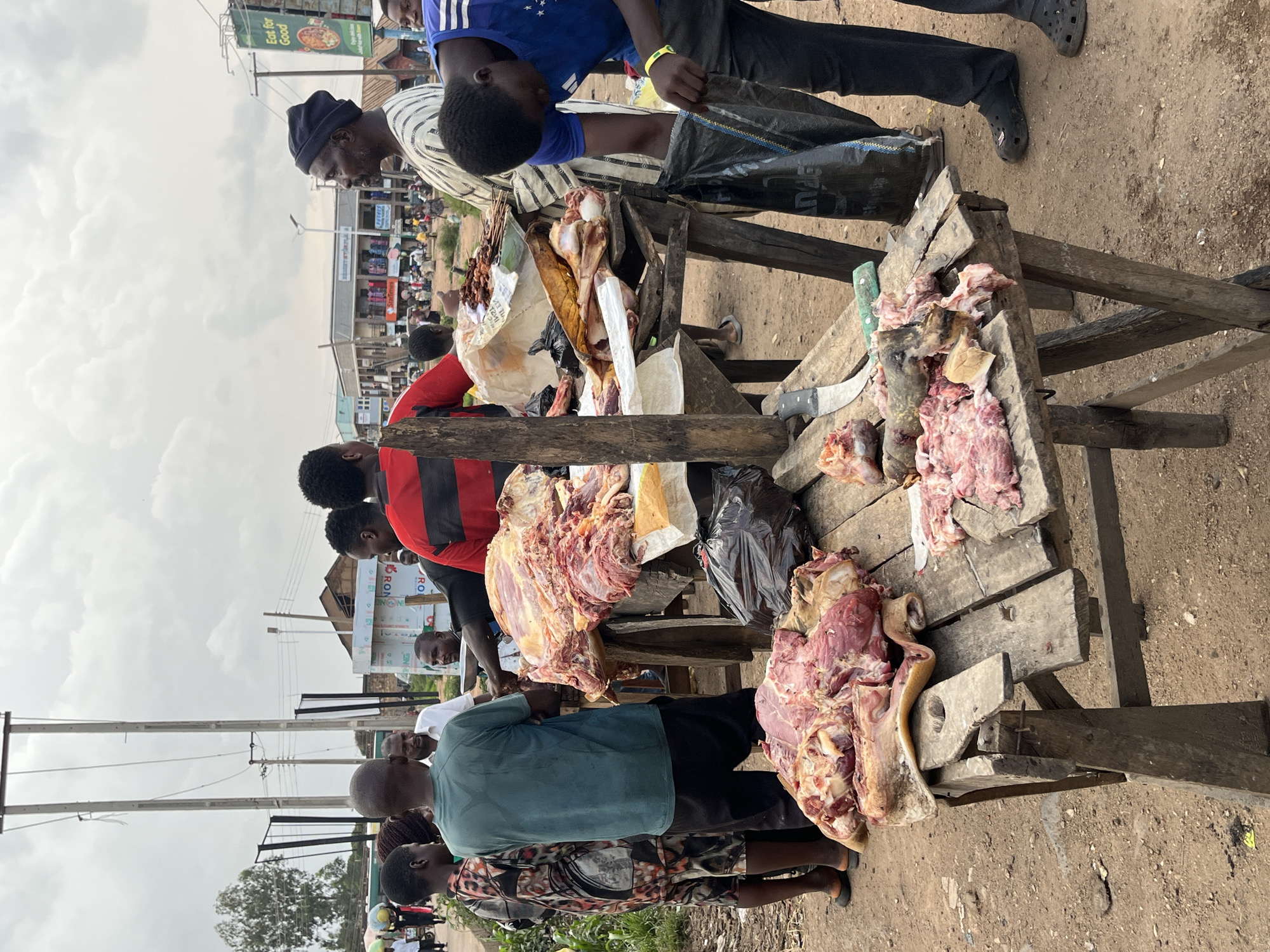 A street market scene is captured in Bukuru, Nigeria, under an overcast sky. The foreground is dominated by two wooden tables laden with raw meat. The larger table on the left displays several large cuts of meat, including what appears to be a pig's head and trotters, alongside other unidentifiable animal parts. The table on the right has smaller portions of meat, some of which have been chopped or prepared for sale. A butcher's knife with a green handle rests on this table, near a larger, older-looking knife. To the right of the tables, a young man in a blue t-shirt and dark pants is seen placing something into a black plastic bag, his face obscured as he looks down.
Several people are gathered around the stalls. On the left, a woman in a patterned dress and a man in a green t-shirt stand facing the meat. Behind them, other individuals are visible, some with their backs to the camera. In the background, a bustling street scene unfolds, with more people, market stalls, and buildings lining the road. Utility poles with extensive wiring are prominent in the middle ground. A sign with the words "Eat for Good" is visible on the right side of the image. The ground is a mixture of dirt and gravel, suggesting an outdoor, unpaved market area. The overall atmosphere appears to be one of commerce and daily activity in a Nigerian urban setting.