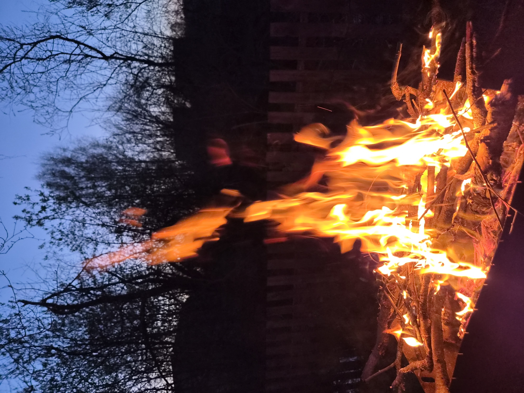 The image shows a bonfire at night, with flames rising from burning wood. To the left of the fire, bare tree branches are silhouetted against a dark blue twilight sky. A dark vertical object, possibly a fence or structure, is visible behind the fire. It is not possible to confirm the location is Узяр, Russia, based solely on the image.