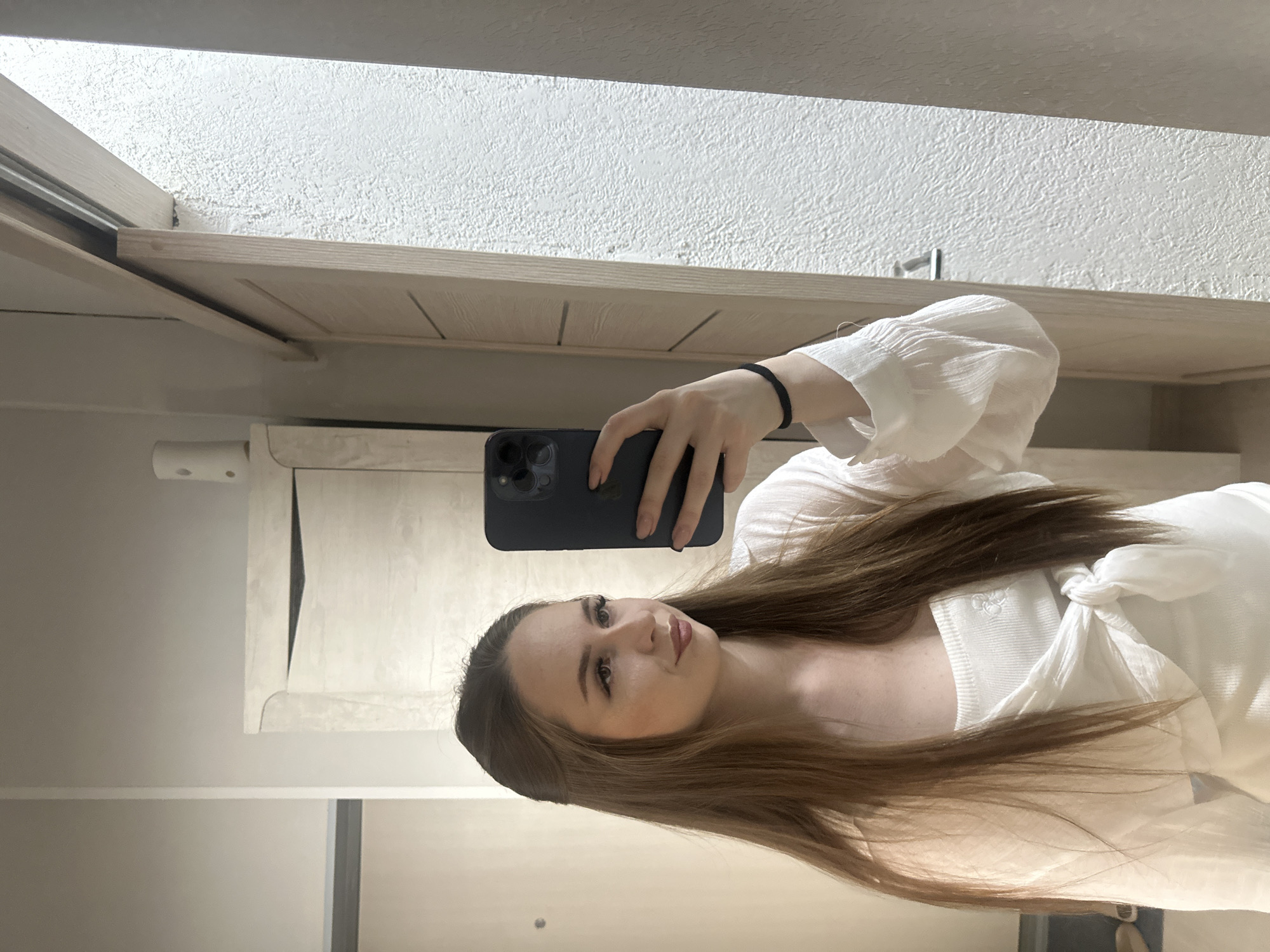 A young woman with long, brown hair is taking a mirror selfie indoors. She is wearing a white top that is tied at the waist, with a sheer white blouse over it. She is holding a dark-colored smartphone in her right hand. The background shows a hallway with light-colored walls and wooden doors. To the right of the woman, a textured wall is visible. The lighting suggests an indoor environment, possibly a home or hotel room.