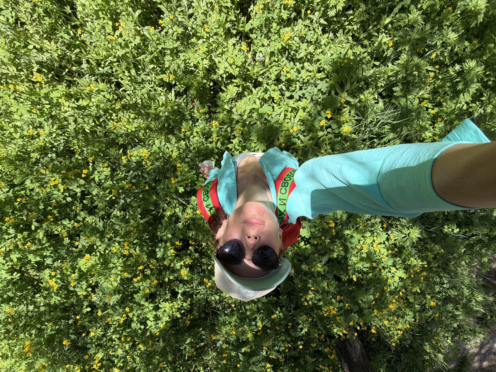 A woman is pictured from a high angle, looking up at the camera while lying or sitting in a field of green grass and small yellow flowers. She is wearing a light-colored baseball cap, sunglasses, and a turquoise garment with red and green accents on the sleeves. The fabric on her sleeves has Cyrillic lettering in white, which appears to read "И СВОБОД" and "ЫХ И СВОБО". The woman's left arm is extended downwards, and she is holding a clear plastic bottle. The background consists of a dense, green ground cover with numerous small yellow flowers, indicative of a natural, possibly park-like setting. The sun is casting shadows, suggesting it is daytime. The image does not contain any discernible elements that specifically identify the location as Birobidzhan, Russia.