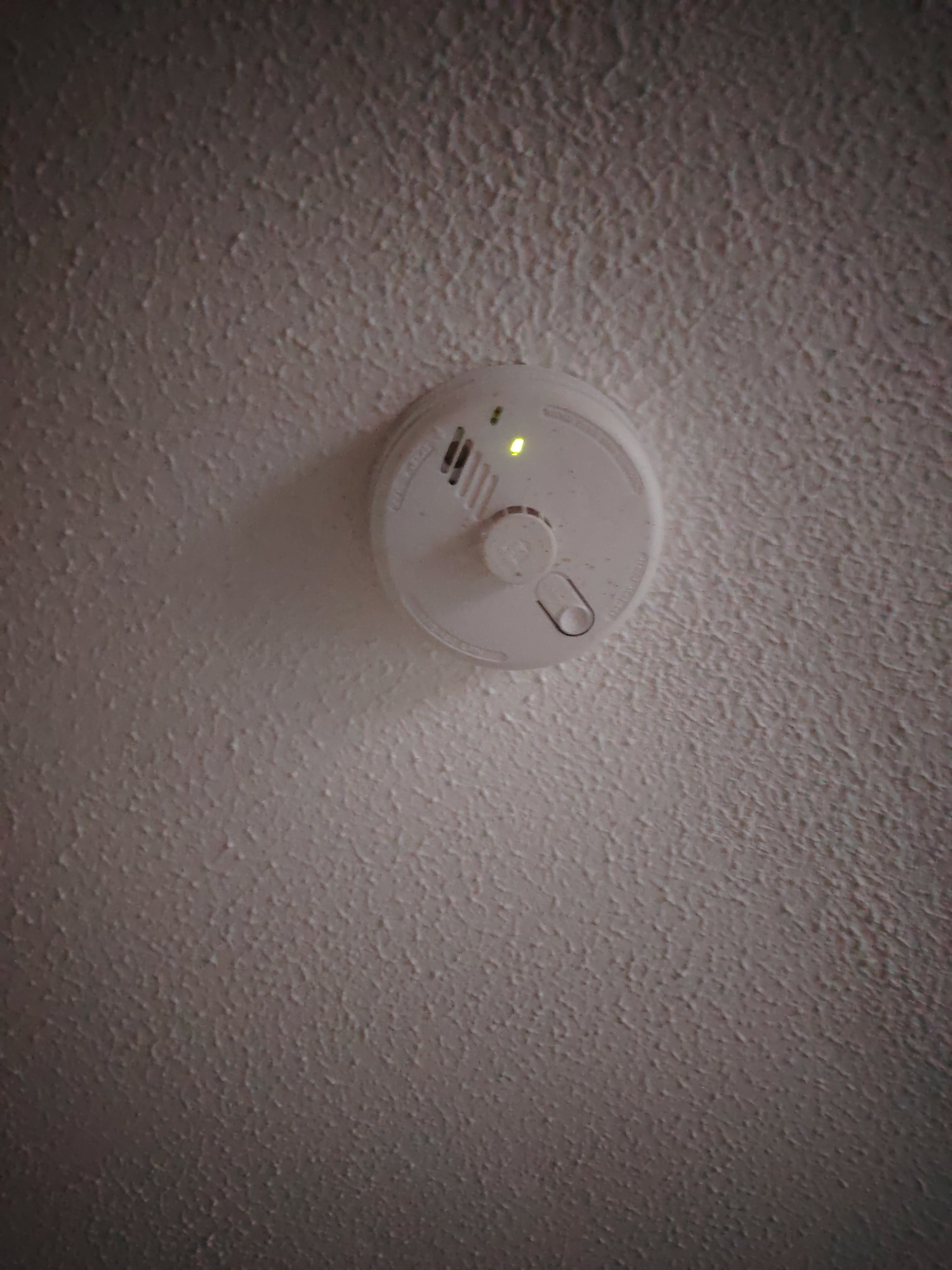 A white, circular smoke detector is affixed to a textured, off-white ceiling. A small, illuminated green LED light is visible on the upper left quadrant of the detector, indicating operational status. The detector features peripheral vents and a central push-button or test switch. A metal tab or hook-like component extends from the lower right side of the device. The ceiling exhibits a rough, stippled, or "popcorn" texture. The scene is moderately lit, with the green LED providing a distinct point of light. The image captures an upward perspective of the ceiling and detector. This scene is located within Huntingdonshire, United Kingdom. No people, other subjects, or actions are depicted.