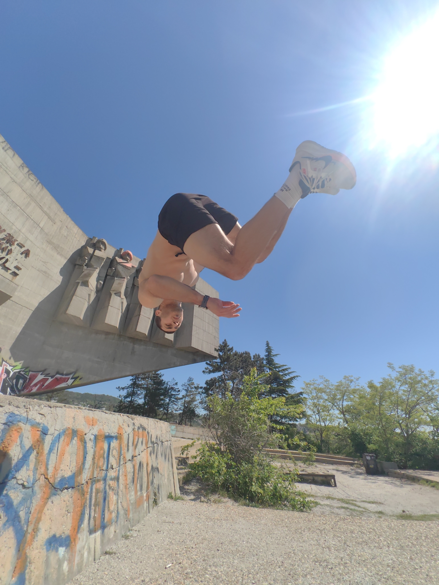 A man is in mid-air performing a backflip in front of the Monument to the Defenders of Stara Zagora in Stara Zagora, Bulgaria. He is wearing black shorts, white socks, and athletic shoes. The sky is bright blue with the sun in the upper right corner. Graffiti covers a concrete wall in the foreground. Trees and a gravel ground are visible in the background.