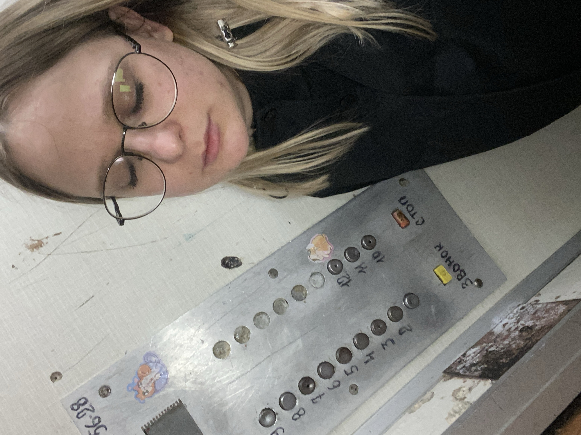 A woman wearing glasses and a dark-colored shirt is lying with her head near an elevator button panel. The panel has Cyrillic text indicating "STOP" and "CALL", along with floor numbers. A sticker is affixed to the lower part of the wall near the elevator panel. There is a label near the sticker containing some of the numbers "56-28". The location is Chaikovsky, Russia.