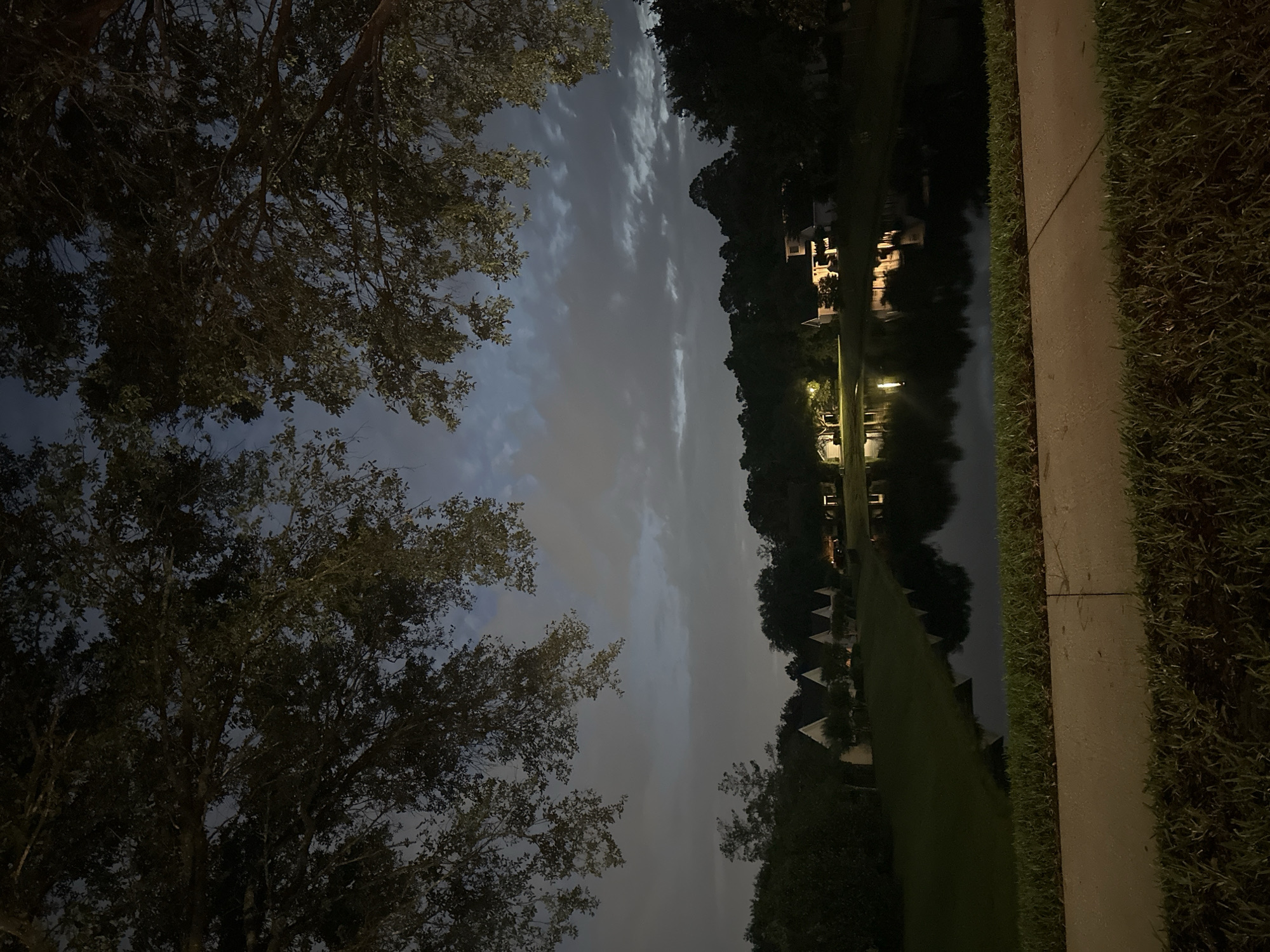 The image, taken at night, shows a sky filled with clouds, framed by trees on the left. A body of water reflects lights from buildings on the right. A sidewalk with grass borders the water. The location is in the city of Jacksonville, United States of America.
