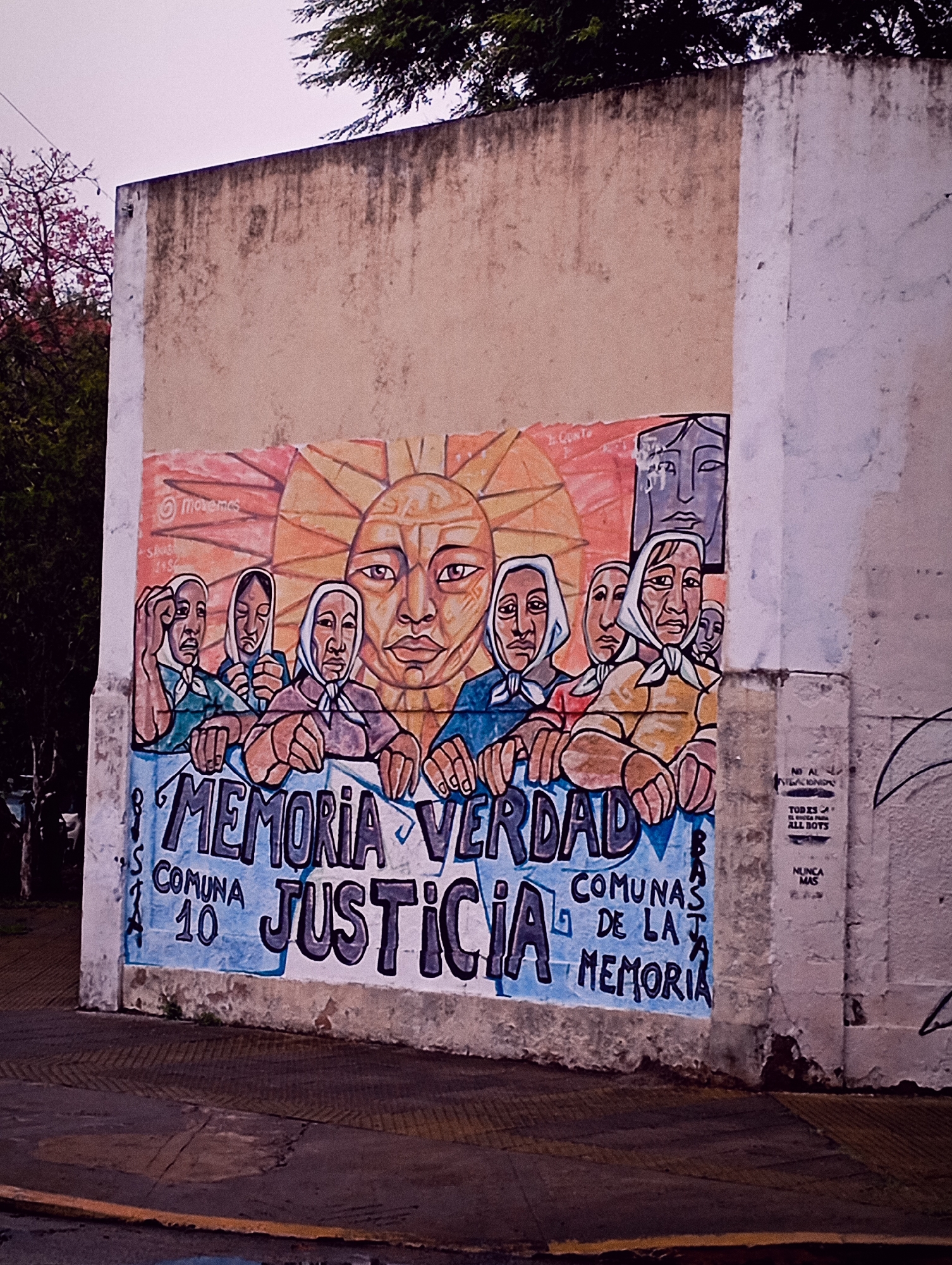 A street-level view captures a painted mural on a weathered building in Buenos Aires, Argentina. The mural, framed by a plain, unpainted wall, depicts a stylized sun with a human-like face above a group of people, presumably women, wearing headscarves. The word "Memoria" (Memory), is prominent alongside "Verdad" (Truth) and "Justicia" (Justice), and the phrase "Comuna 10 de la Memoria" (Commune 10 of Memory). The style is somewhat faded and rough. To the right of the mural, on a white wall, are smaller painted messages and graffiti. The street and sidewalk in the foreground suggest an urban setting, with trees and foliage visible in the background. The sky is overcast.