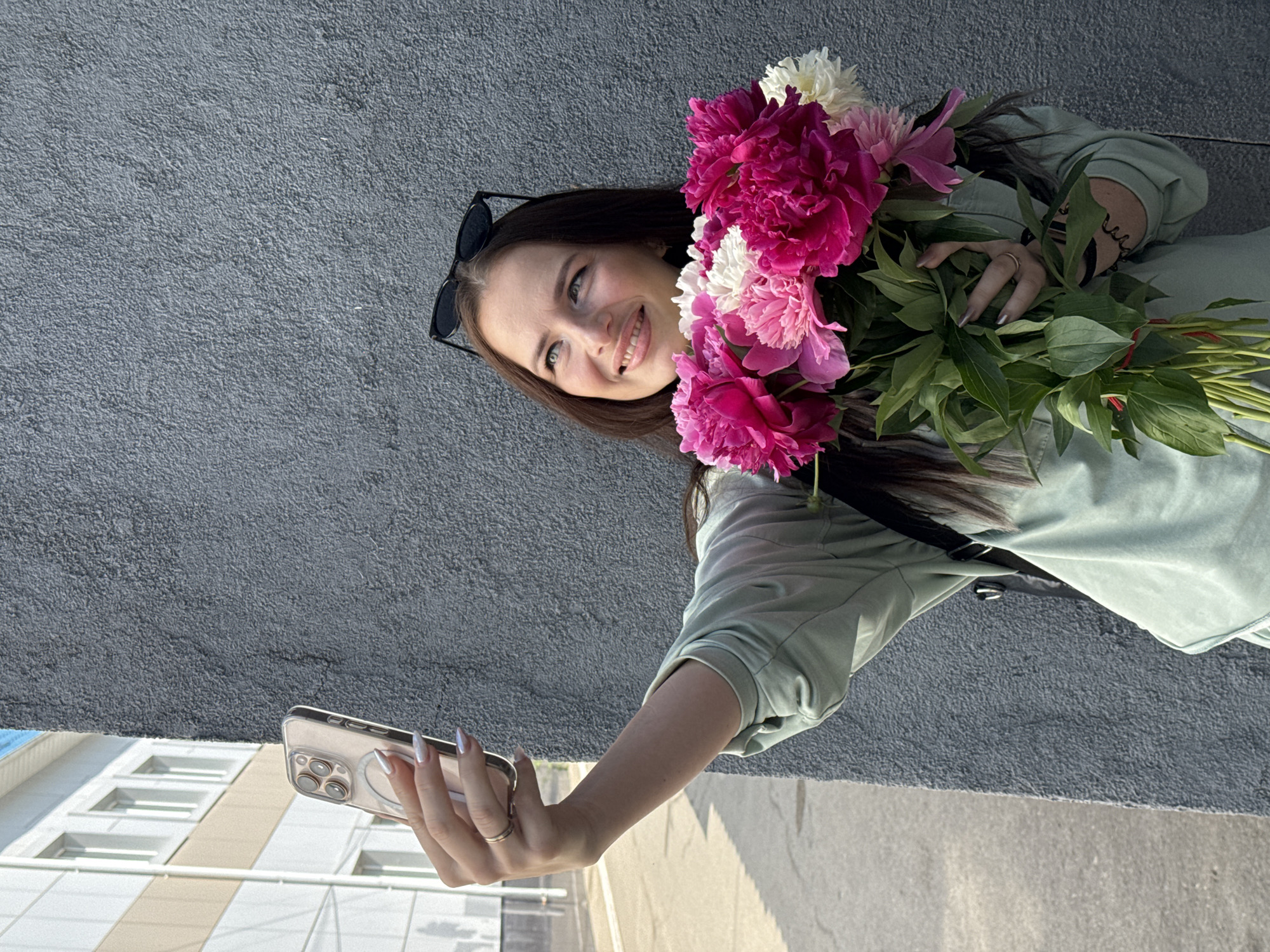 A young woman with long dark hair and sunglasses perched on her head is taking a selfie with a bouquet of pink and white peonies. She is wearing a light green sweatshirt and a ring on her left ring finger. The bouquet is held in front of her, obscuring part of her torso. She is standing against a textured gray wall, with a building visible in the background. The scene appears to be outdoors, with sunlight casting shadows. This location is likely in the city of Chaikovsky, Russia, given the architectural style of the building and the overall atmosphere.