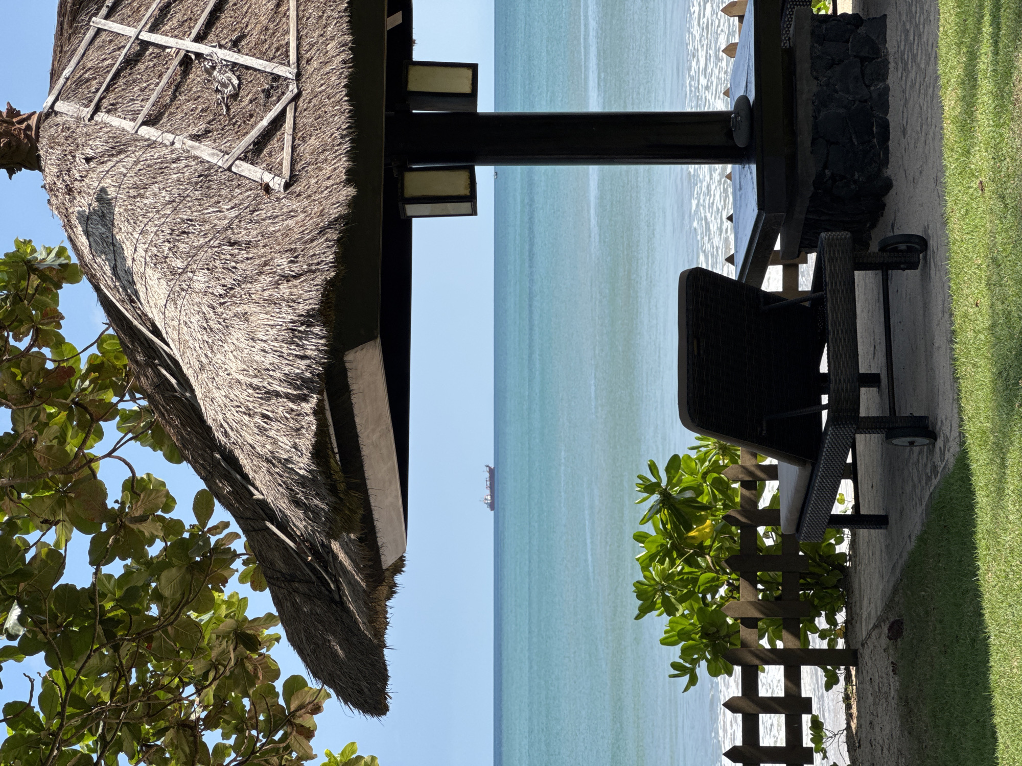 The image, taken in Kuta Selatan, Indonesia, presents a scene overlooking the ocean. A thatched roof structure, possibly a beach hut or gazebo, frames the left side of the image, with foliage partially obscuring it. A black table and chair set are situated on a concrete surface on the right, near a patch of green grass and a short wooden fence with surrounding foliage. In the distance, a ship is visible on the horizon, punctuating the expanse of blue water and clear sky.