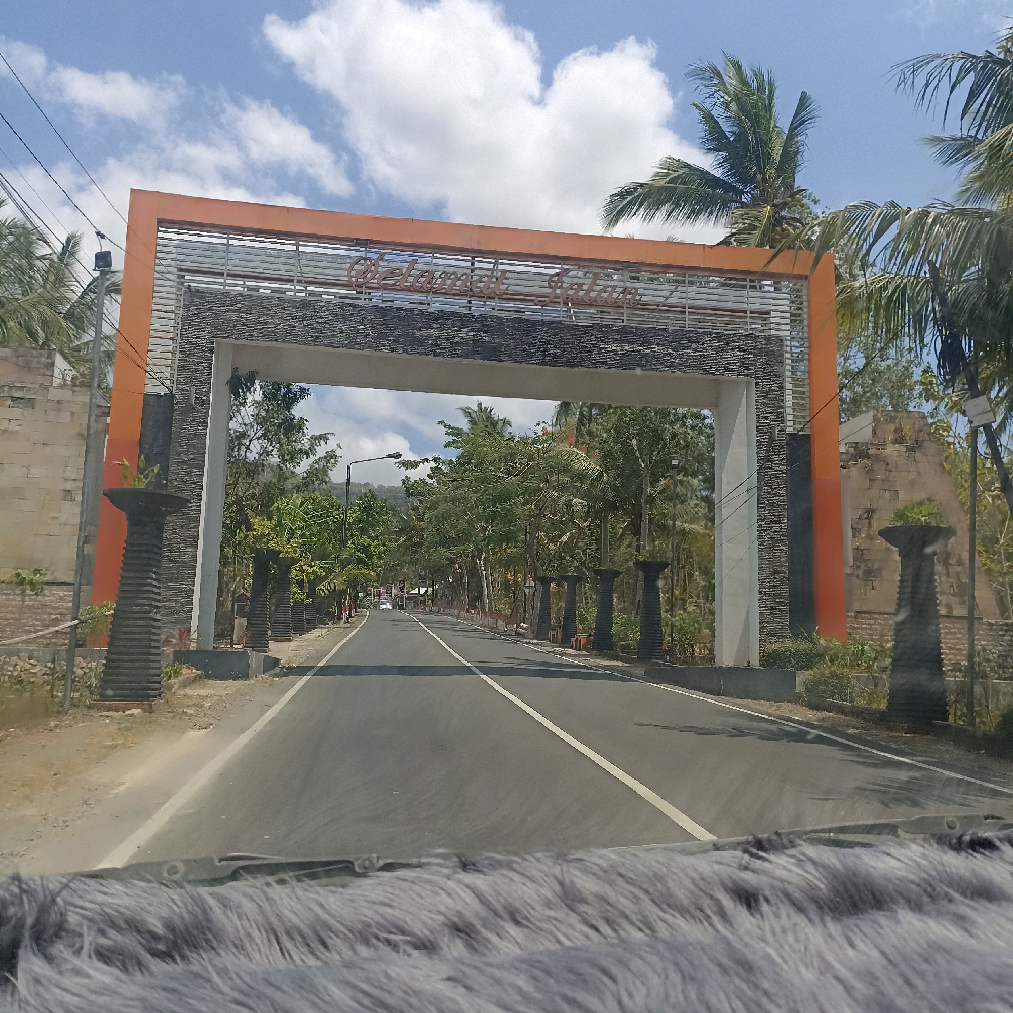 The image captures a road leading through an orange and stone archway in Sedeng, Indonesia. The archway is adorned with the text "Selamat Datang", which translates to "Welcome" in Indonesian. Palm trees are visible on either side of the road, suggesting a tropical or subtropical climate. The sky is blue with scattered white clouds. The road is paved and appears to be a main thoroughfare, with a white line dividing the lanes. The archway is flanked by decorative pillars, some of which hold potted plants. On the right side of the road, beyond the archway, there is a structure made of weathered stone blocks. The foreground of the image shows a blurred view of a car's dashboard and windshield wiper, indicating the photo was taken from inside a vehicle. The overall scene depicts a welcoming entrance to a town or area.