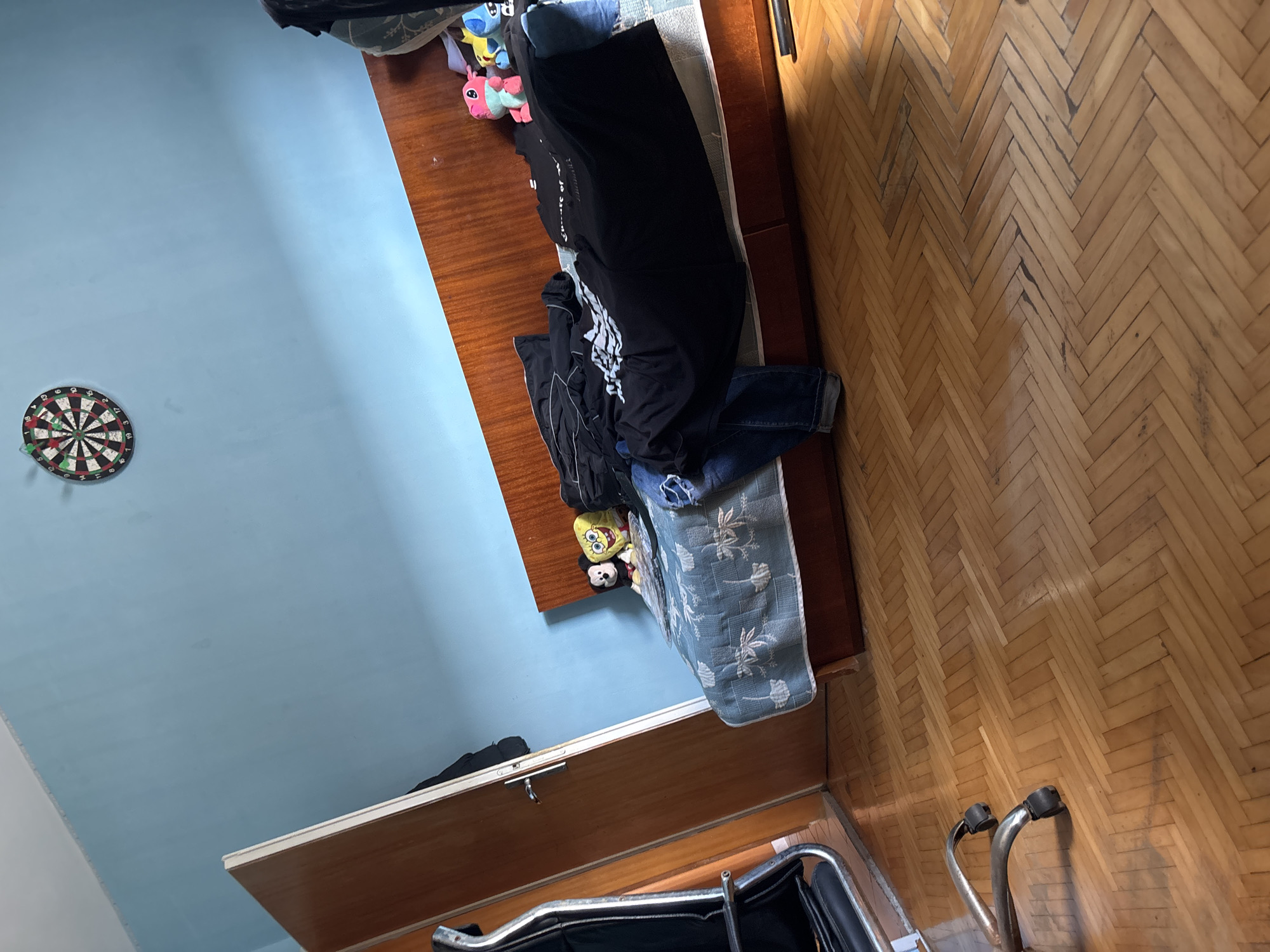 This image depicts an indoor room, likely in Pleven, Bulgaria, characterized by light blue walls and a wooden parquet floor laid in a herringbone pattern. The floor shows signs of wear and tear with some darker areas.
A wooden bed frame or sofa base is positioned against one of the blue walls, topped with a light blue mattress or cover featuring a subtle white floral or abstract design. Clothes are casually piled on this surface, including a black long-sleeved item, a black t-shirt with partially visible white text that reads "Sports of...", and a pair of blue jeans. Near the head of the bed, several plush toys are visible: Spongebob Squarepants, a black and white panda-like toy, and two colorful alien-like characters, resembling Stitch and Angel from Lilo & Stitch.
Mounted on the light blue wall is a dartboard with several darts stuck in it, indicating a recreational element in the room. In the lower part of the image, a wooden piece of furniture, possibly a low cabinet or wardrobe, features a white top edge and a metallic latch. The foreground and bottom right corner reveal components of what appears to be a wheelchair or similar chair, with curved metallic tubing and black armrests or grips.
The room appears to be a lived-in, personal space. There are no people visible, and no specific activity is captured. The lighting suggests an indoor setting, likely with natural light, casting some shadows on the floor.