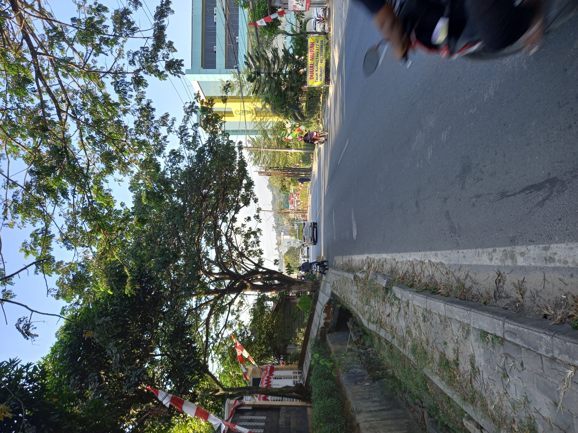 A daytime, eye-level outdoor shot depicts a street scene in Samarinda, Indonesia. The foreground shows a cracked and overgrown concrete sidewalk adjacent to an asphalt road. Red and white flags are visible on poles along the sidewalk, suggesting a possible celebration or event. Several trees with lush green foliage line the street, with their branches extending over the road.
In the background, a blue and light yellow multi-story building stands prominently, likely an office or educational institution. A yellow banner with Indonesian text is displayed on the side of the road, which appears to advertise a sale. A few vehicles, including cars and motorcycles, are present on the road. One motorcycle rider in a red helmet is visible approaching the viewer. Another motorcycle's front portion and a person's hand are blurred in the lower right corner, indicating motion. The sky is clear and blue. The scene appears to be a typical urban environment with a mix of residential and commercial elements.