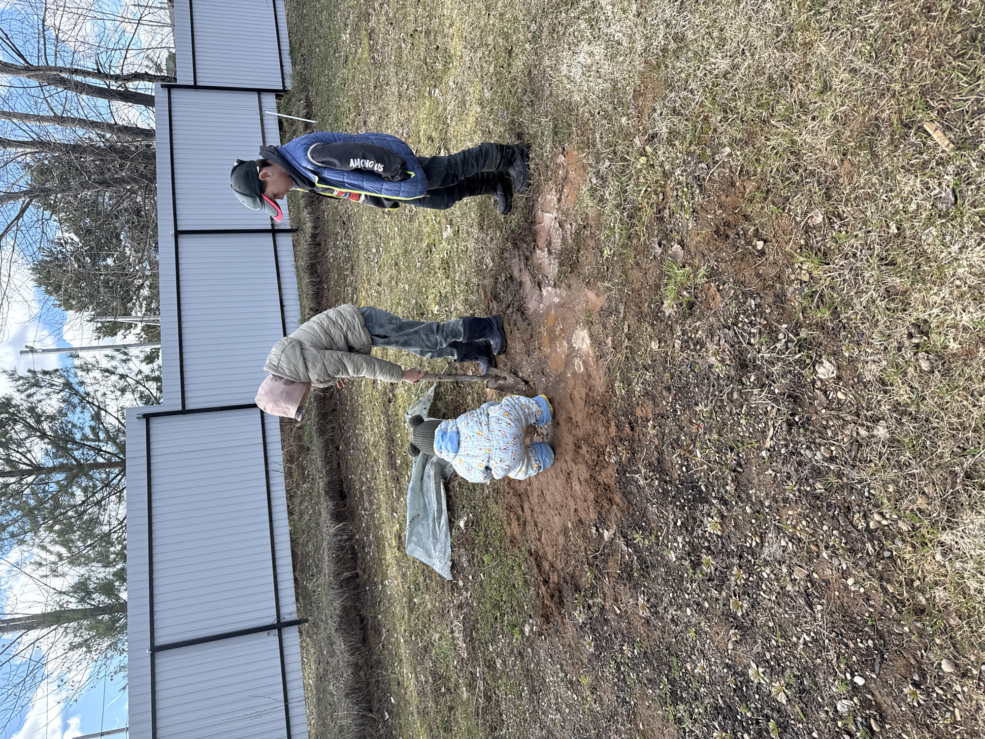 The image shows three individuals outdoors near a fence in what appears to be a yard or garden area in Ольховка, Russia. One person is digging in the dirt with a shovel, while another stands nearby observing. A small child in a patterned snowsuit is crouching near the freshly dug area. The environment appears to be transitioning between seasons, with brown grass and some bare trees in the background. The fence is made of light-colored panels with dark frames. The clothing of the people suggests cool weather.