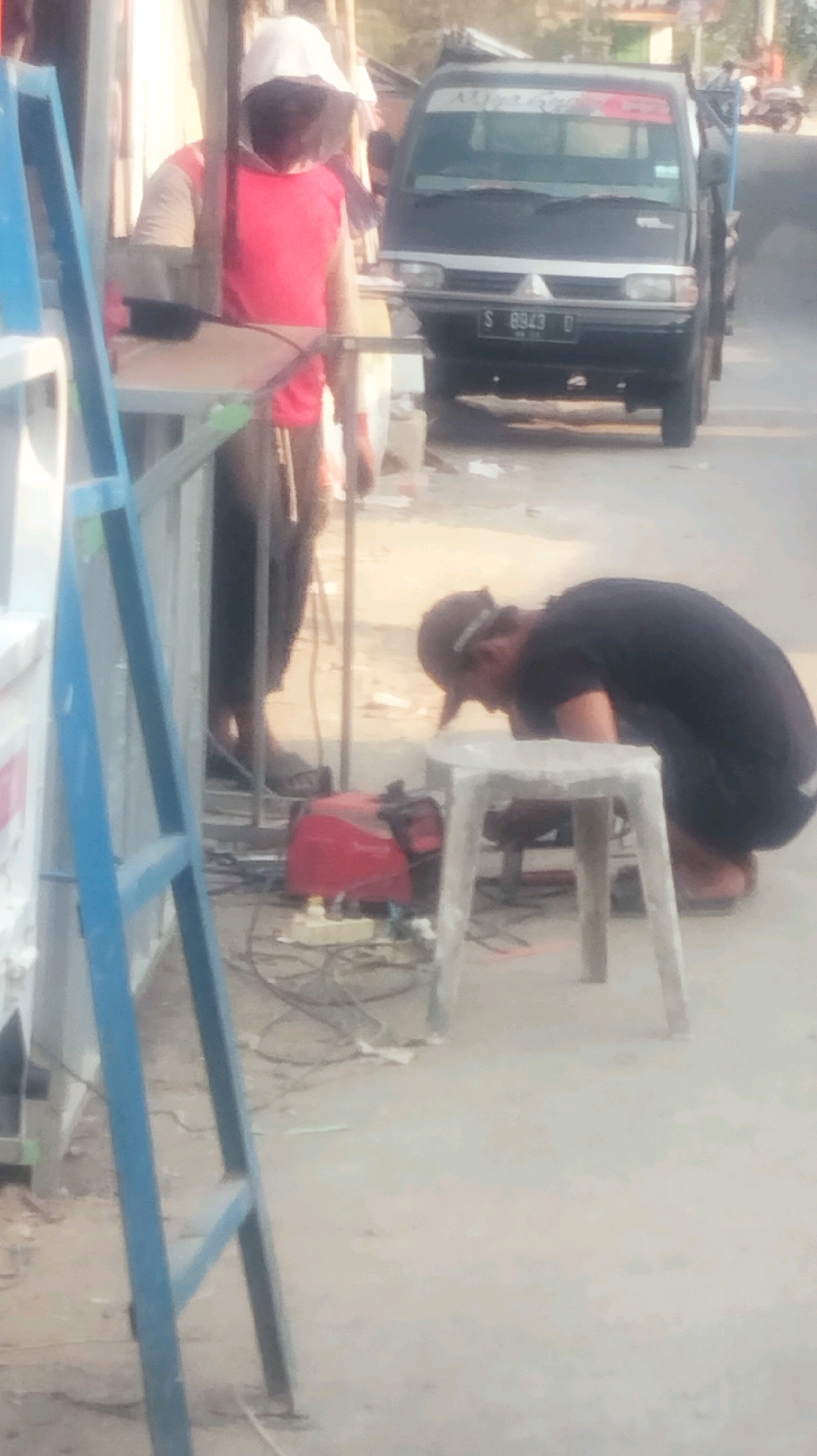The image depicts an outdoor scene on a street in Palang, Indonesia. In the foreground, a person wearing a black t-shirt and a backward baseball cap is bent over a small, white, stool-like object, appearing to be working on something. To the left, a blue ladder is positioned vertically. Behind the kneeling person, a red welding machine and other electrical equipment are visible on the ground. Further back, another person wearing a white hat and a red and tan shirt stands near a metal frame. A black pickup truck with a license plate reading "S 8343 D" is parked on the right side of the street. The environment appears to be a roadside with a paved surface.
