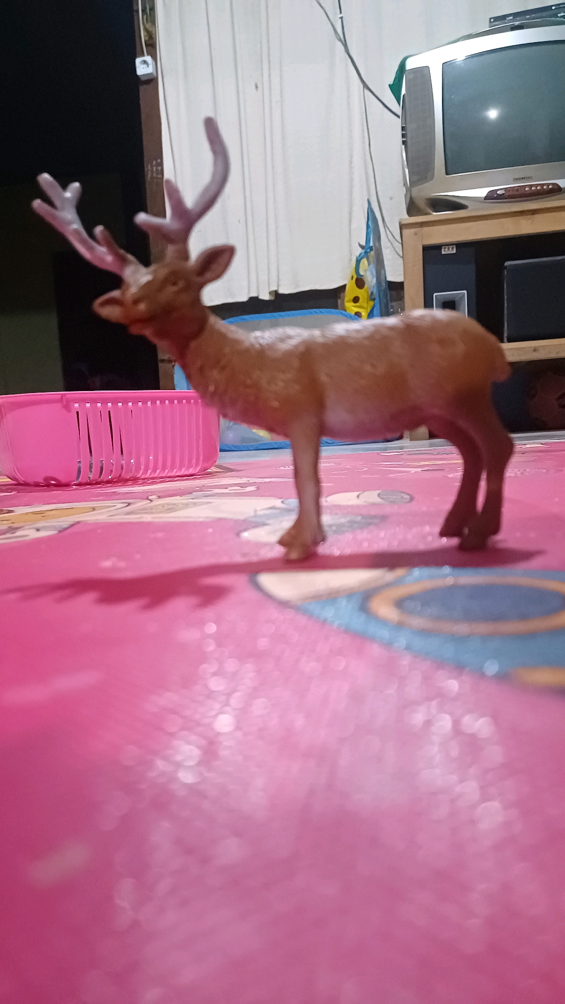 The image shows a plastic toy deer standing on a pink mat. The deer is brown and has antlers. In the background, there is a pink basket, a television set, and white curtains. The scene appears to be indoors, possibly in a living room. The location is not specified to be Wonokerso, Indonesia.