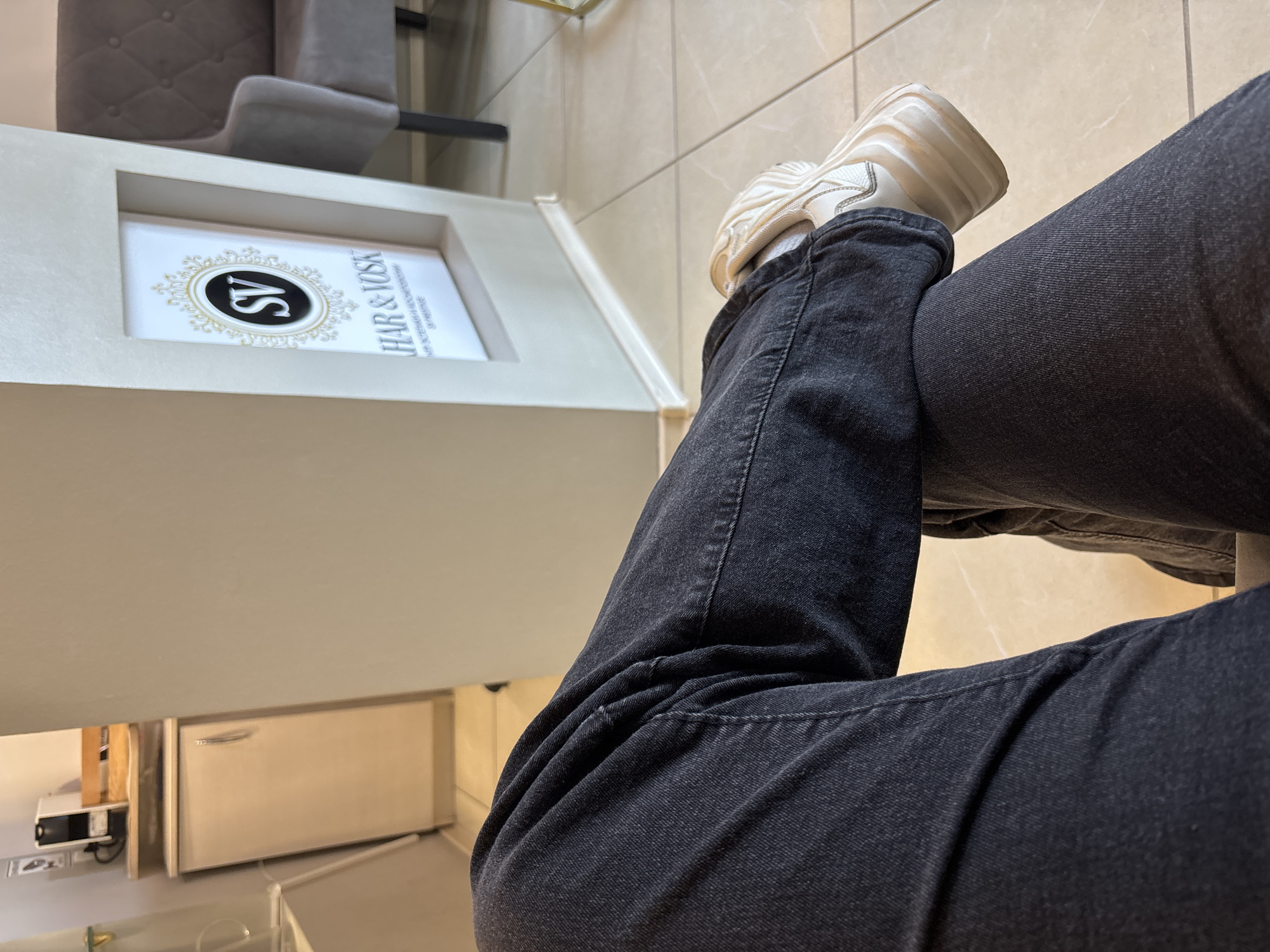 The image shows a person's legs and feet, clad in dark wash jeans and white sneakers, crossed at the ankles. The background suggests an indoor setting, possibly a waiting area or salon. To the left, a cabinet and a coffee machine are visible on a shelf. In the center, a pillar displays a sign with "SV" in a circular emblem above the text "HAIR & VOSK" and smaller, less legible text. To the right, a portion of a tufted grey armchair is visible, and the floor is tiled. The overall impression is one of a clean, modern interior. The location is identified as Unknown, Russia, but no specific urban details are discernible from the image.